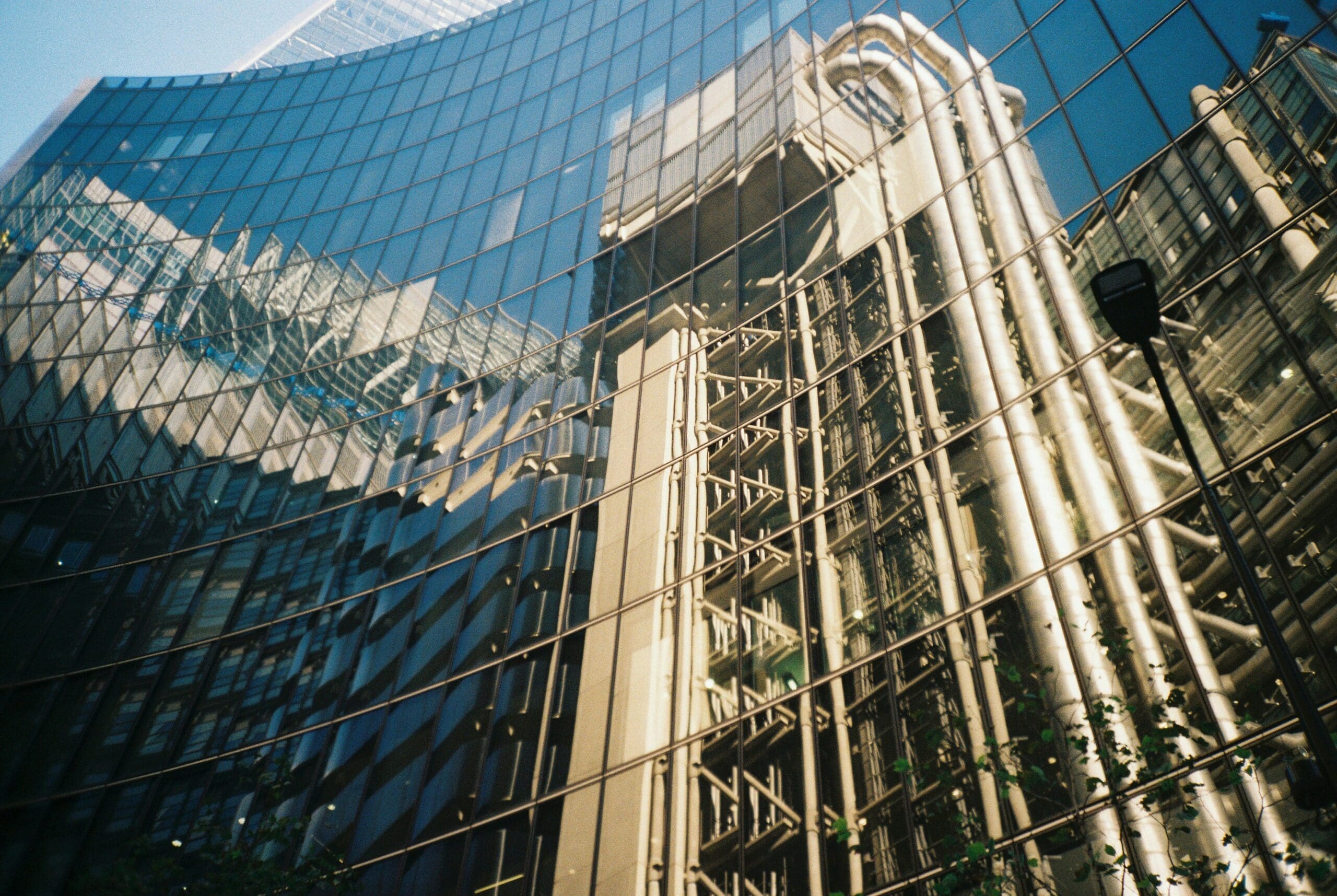 Un bâtiment moderne en verre reflète l'image d'un autre édifice aux tuyaux apparents et aux structures industrielles, créant ainsi un effet déformé et abstrait sous un ciel bleu limpide.