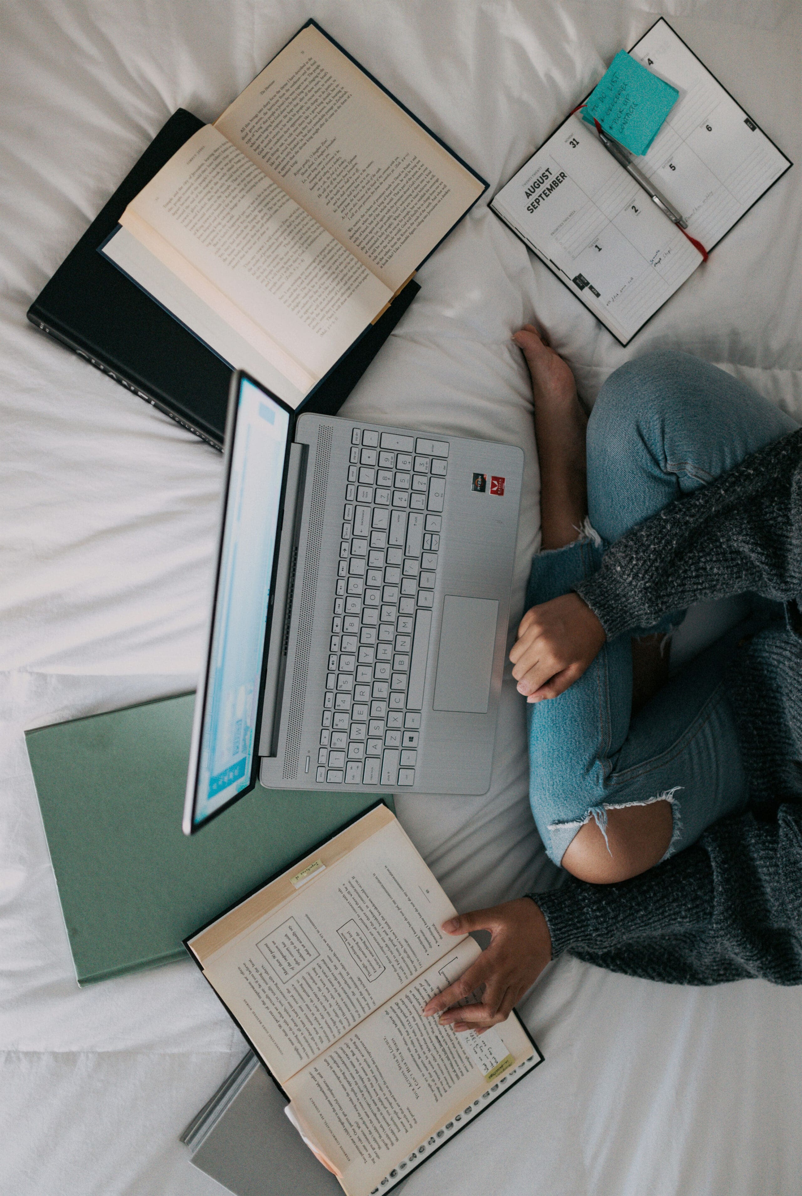 Une personne est assise en tailleur sur un lit, entourée d'un ordinateur portable, de livres ouverts, d'un cahier et d'un agenda, et semble étudier ou travailler dans un cadre chaleureux.