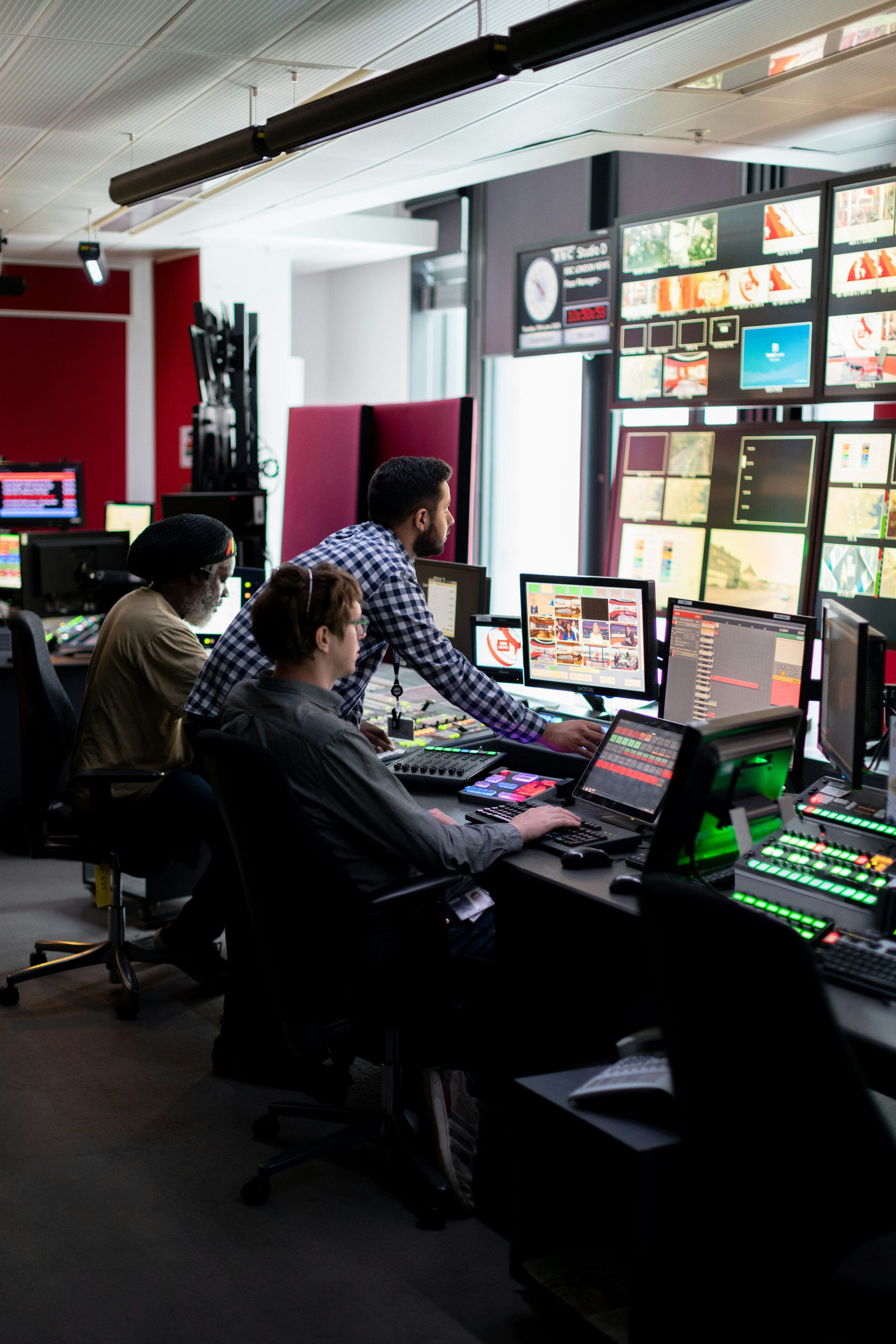 Trois personnes travaillent dans une salle de contrôle moderne, où elles surveillent plusieurs écrans affichant des flux vidéo et des graphiques, entourées d'ordinateurs et de panneaux de commande.