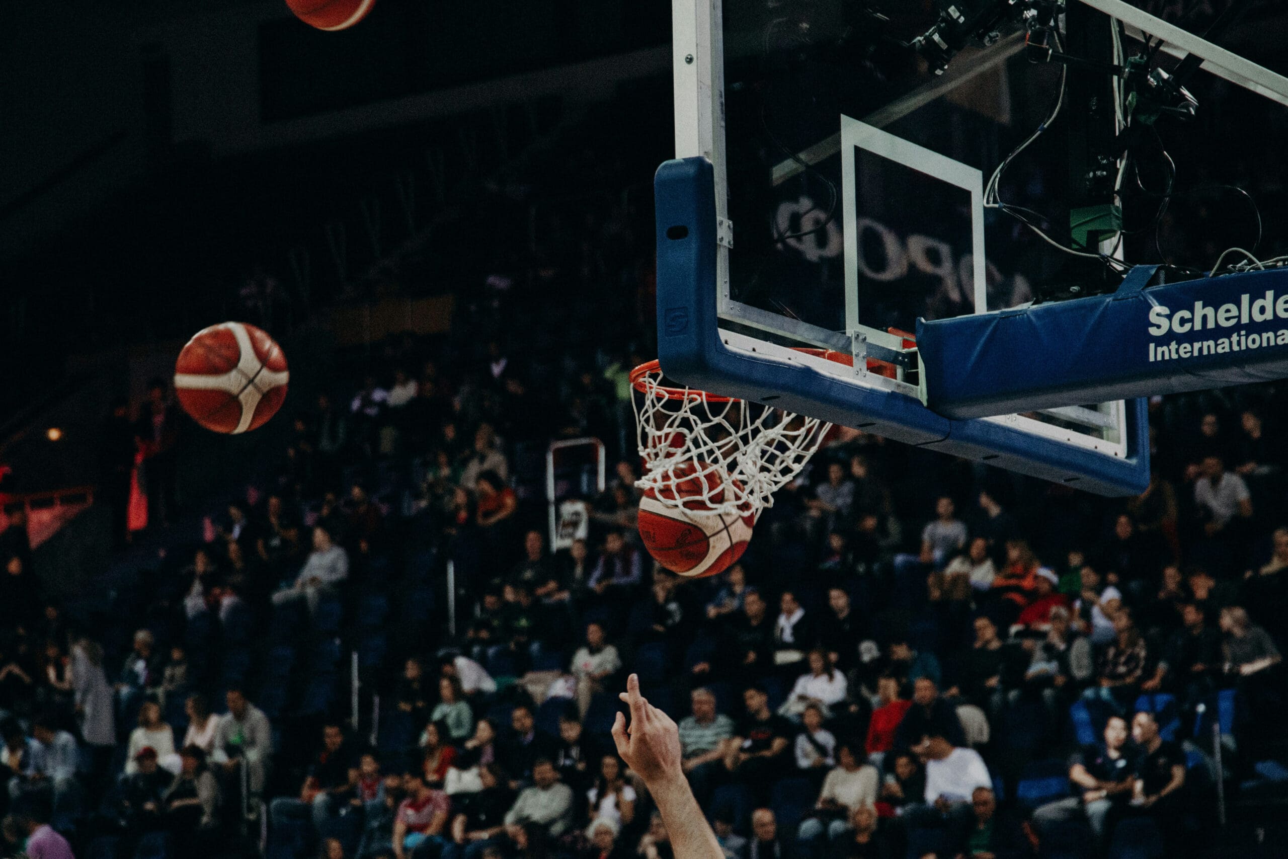 Un ballon de basket sur le point de passer dans le panier pendant un match, avec la main d'un joueur visible et une foule de spectateurs floue en arrière-plan.