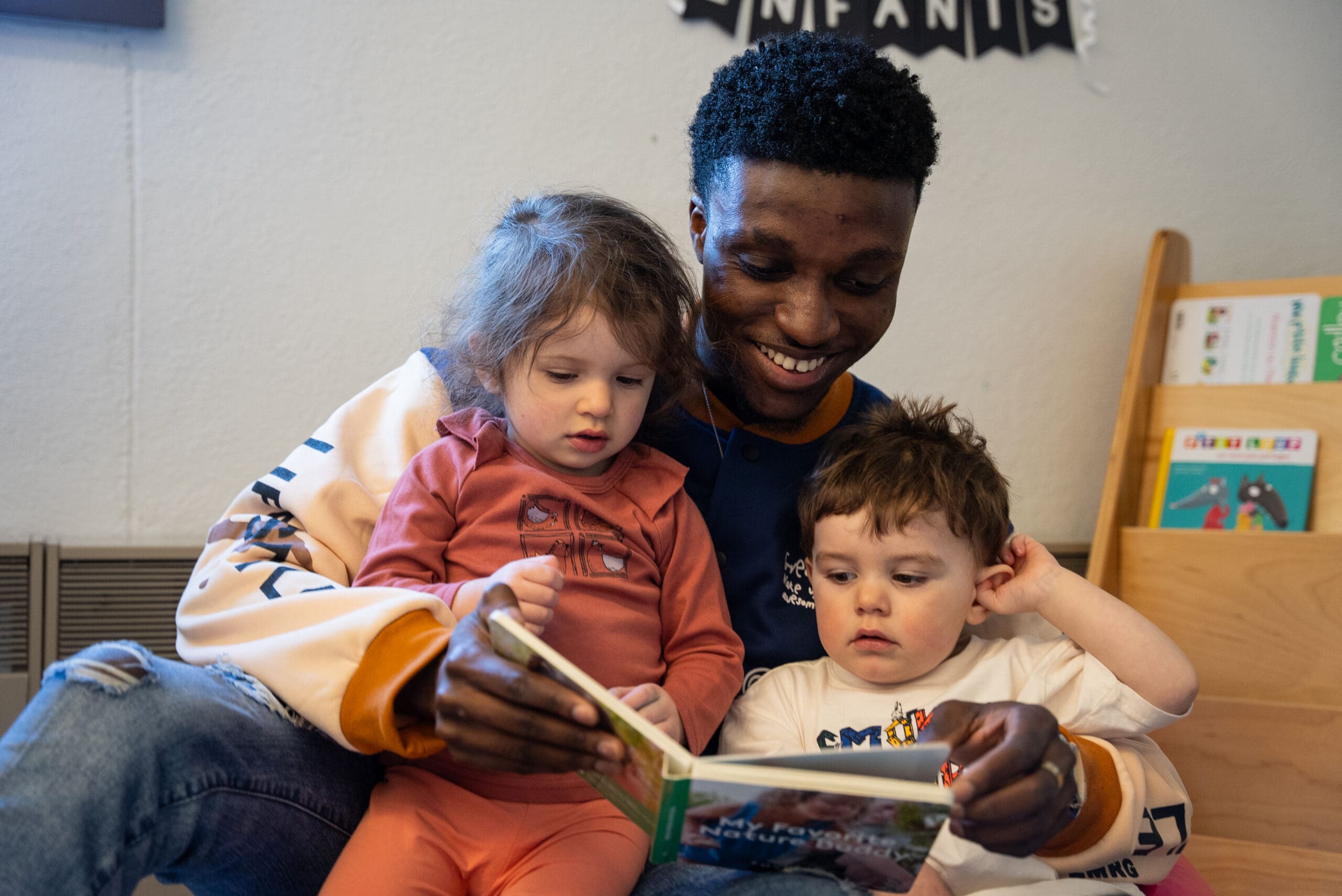 Un homme souriant lit un livre à deux jeunes enfants assis sur ses genoux, une fille et un garçon, dans une pièce chaleureuse où l'on aperçoit, en arrière-plan, une bibliothèque et une banderole pour enfants.