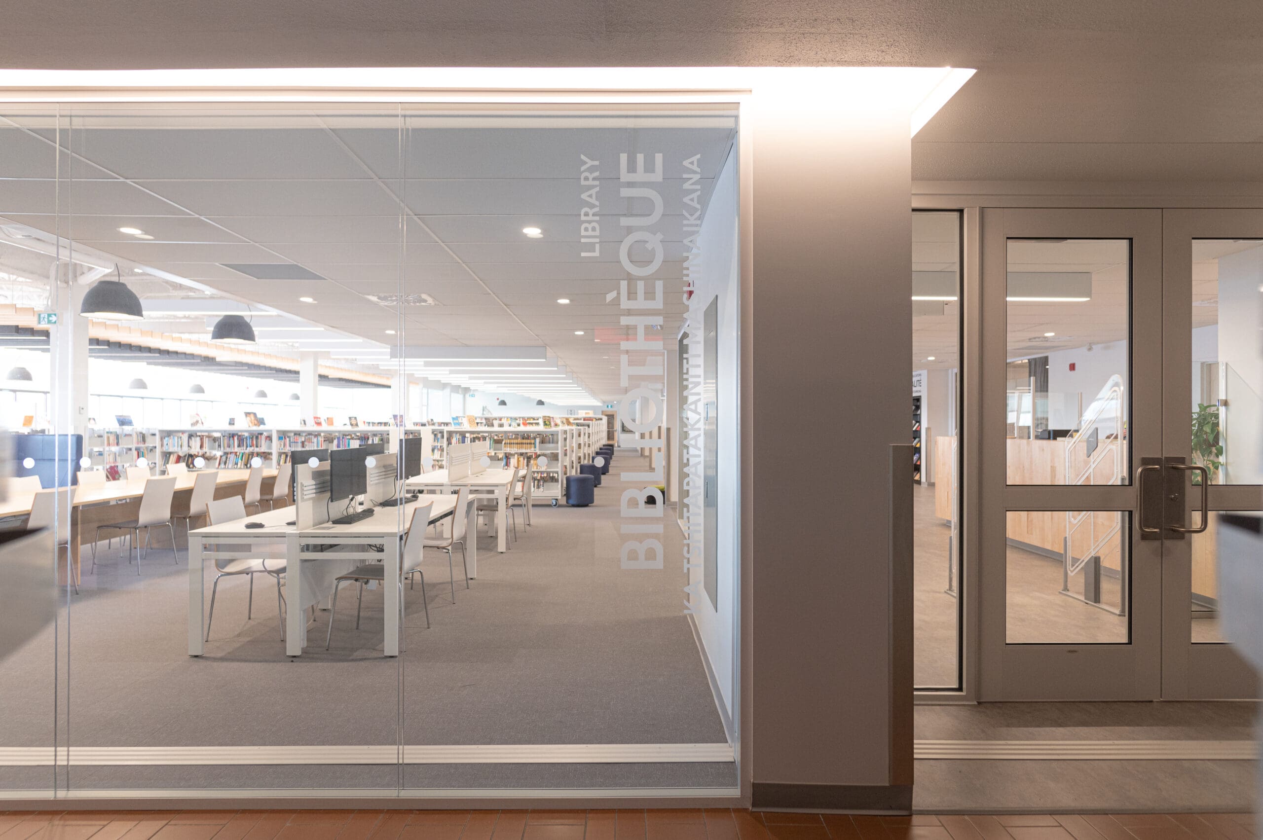 Entrée de la bibliothèque aux parois vitrées, ornée des inscriptions « BIBLIOTHÈQUE » et « LIBRARY » en lettres blanches. À l'intérieur, on aperçoit des rangées de tables, de chaises et d'étagères dans un espace lumineux et moderne doté de grandes baies vitrées.