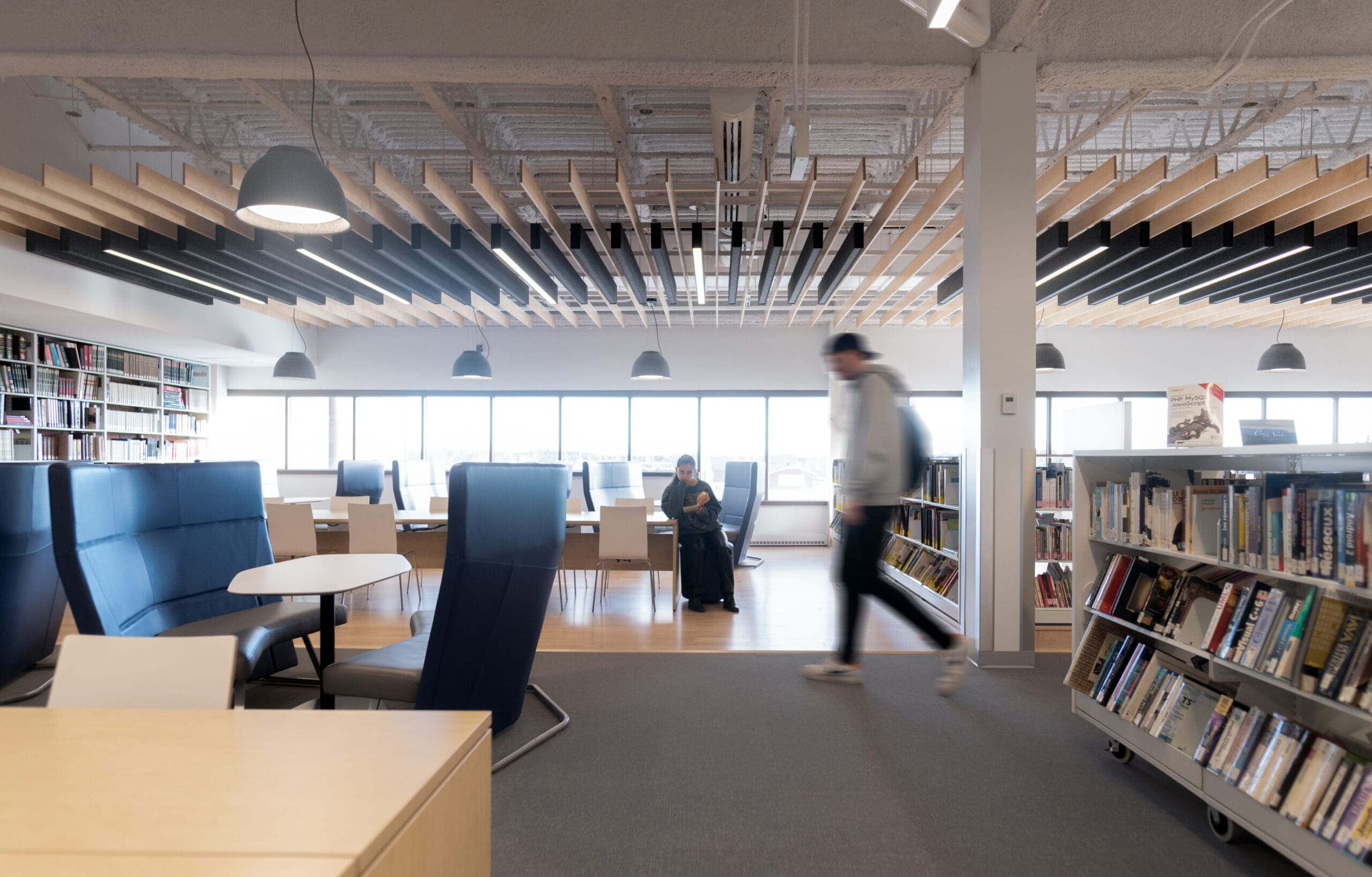 Une personne est assise et lit près de grandes baies vitrées dans une bibliothèque moderne, tandis qu'une autre personne passe à l'arrière-plan, floue, entourée d'étagères et d'un mobilier contemporain.