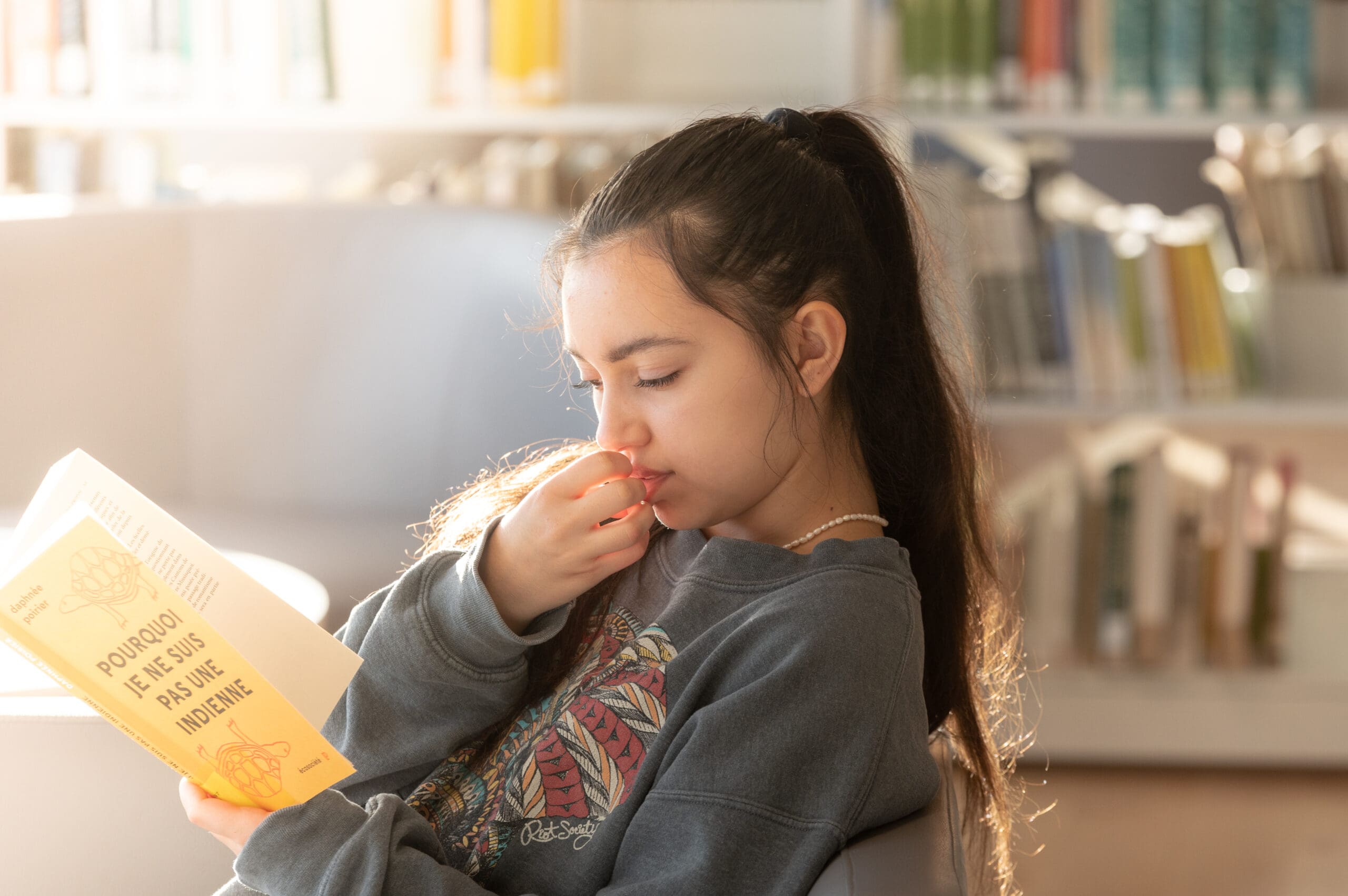Une jeune femme aux longs cheveux bruns est assise dans une bibliothèque, plongée dans la lecture d'un livre jauni intitulé « Pourquoi je ne suis pas indien ». La lumière du soleil illumine son visage, tandis qu'en arrière-plan, les rayonnages de livres apparaissent flous.