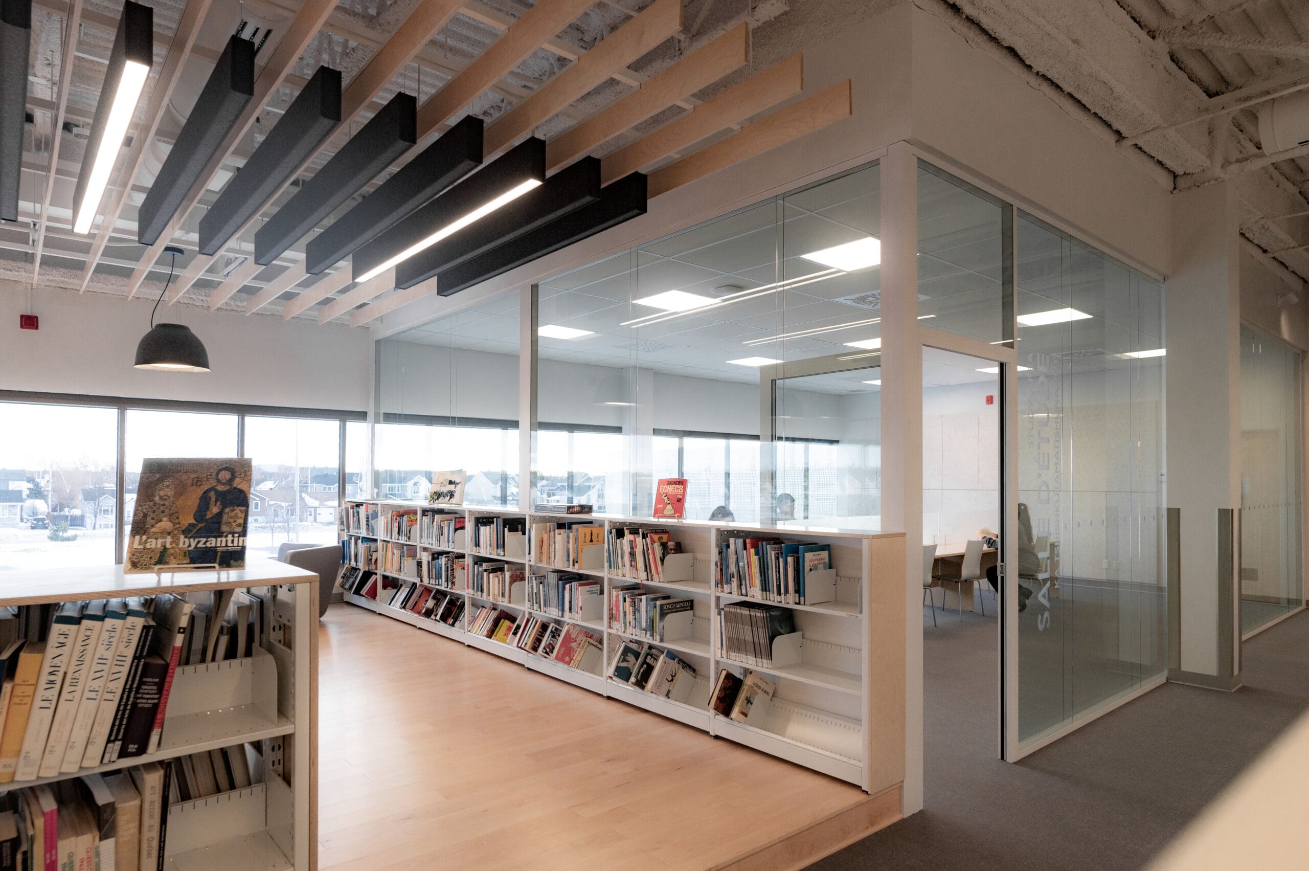 Un espace de bibliothèque moderne composé d'étagères blanches basses regorgeant de livres, d'un parquet en bois clair et d'une salle d'étude vitrée. Au plafond, des panneaux acoustiques originaux sont suspendus sous une grille de planches en bois clair, avec une suspension noire à proximité.