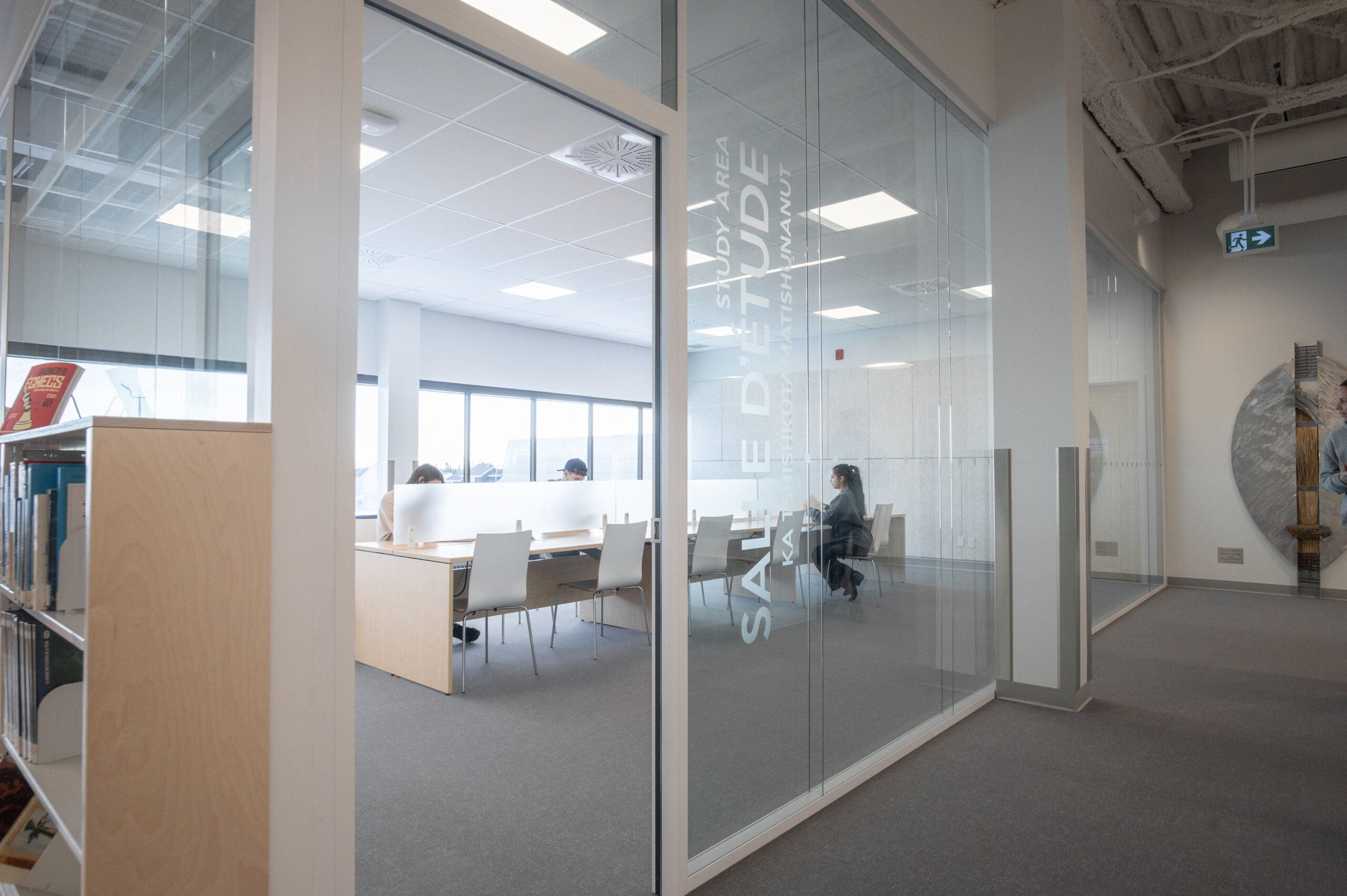 Une salle d'étude moderne aux parois vitrées, où plusieurs personnes sont assises à des tables et où l'on aperçoit des étagères depuis l'extérieur. La lumière naturelle inonde la pièce.