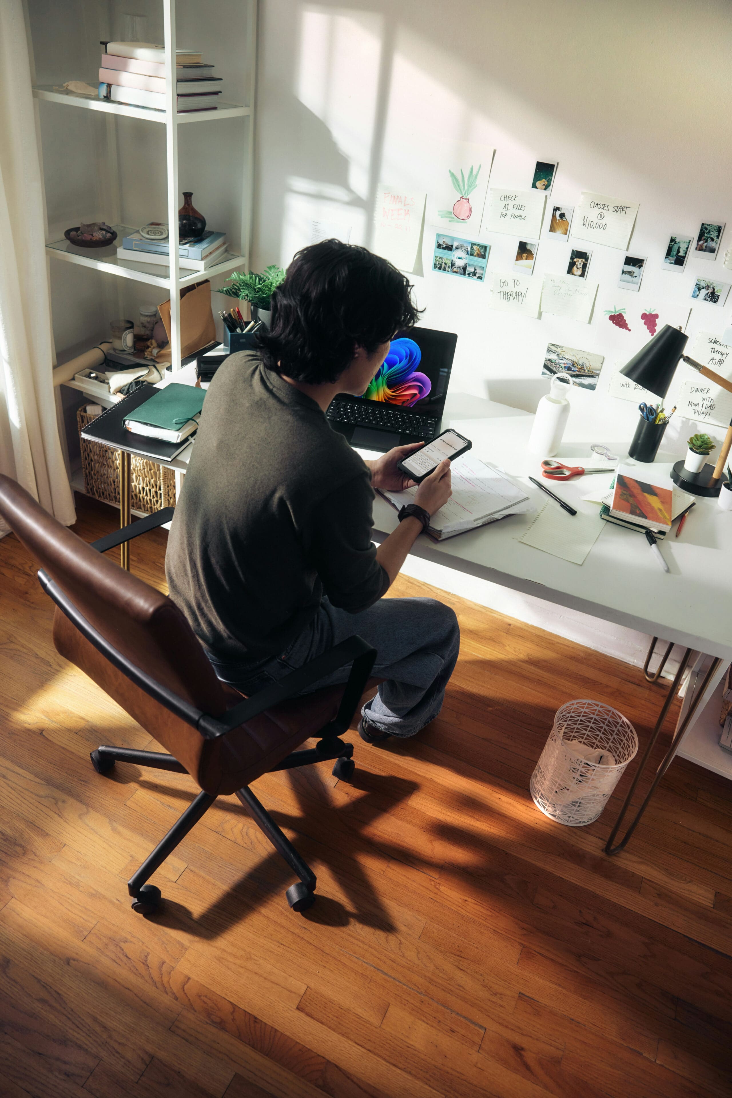 Une personne est assise à un bureau blanc, un smartphone à la main, entourée d'un ordinateur portable, de cahiers, d'une lampe et de matériel de dessin. L'espace de travail est bien rangé, avec des étagères, des notes et des photos accrochées au mur, et la lumière du soleil qui inonde la pièce.
