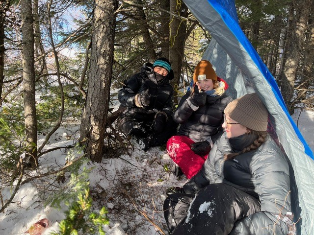 Trois personnes vêtues de tenues d'hiver sont assises sous une bâche bleue dans une forêt enneigée.
