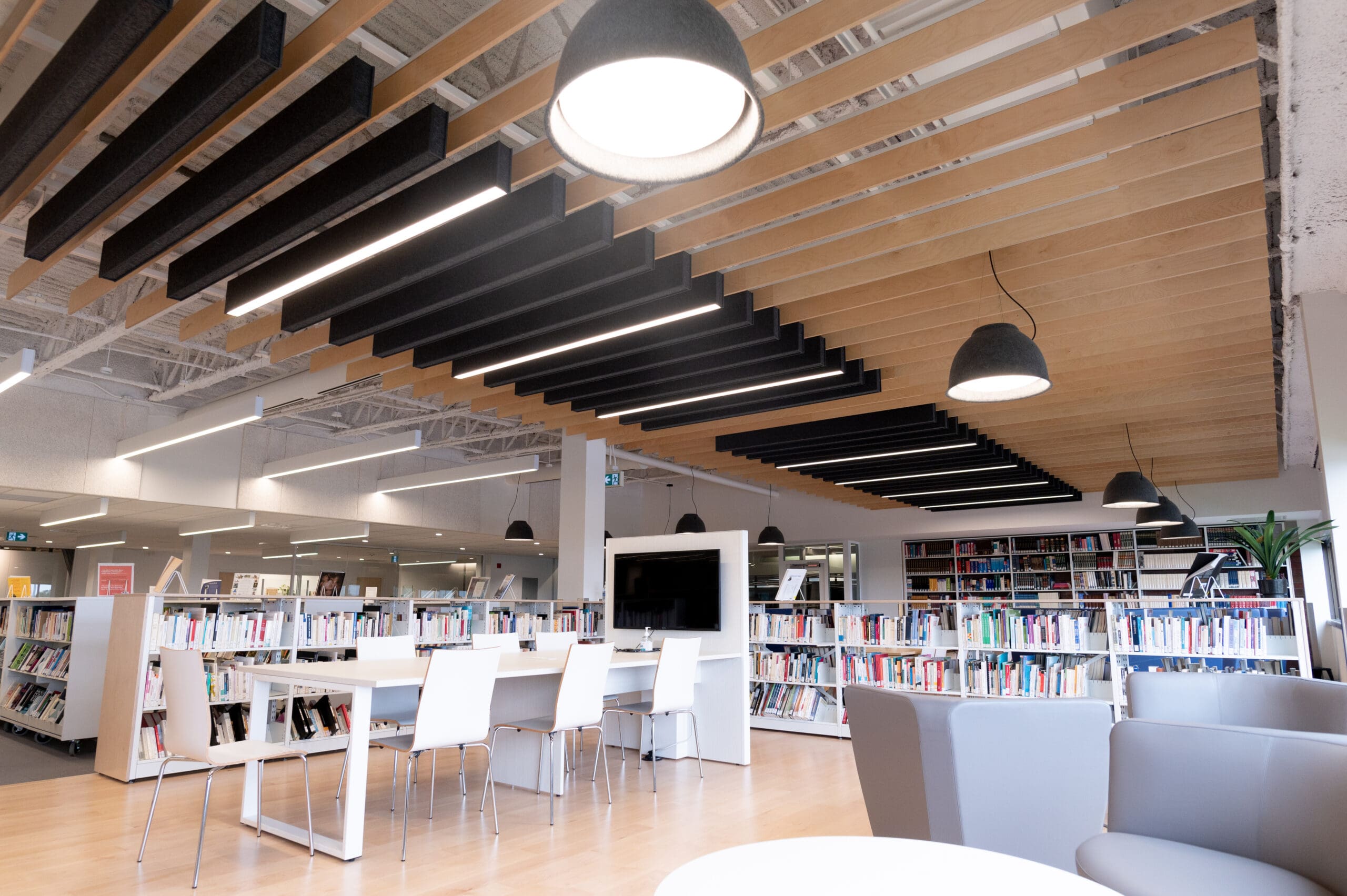 Intérieur de bibliothèque moderne avec des tables et des chaises blanches, des étagères remplies de livres et de grandes suspensions noires suspendues à un plafond à lattes de bois. On aperçoit également, au premier plan, quelques fauteuils gris.
