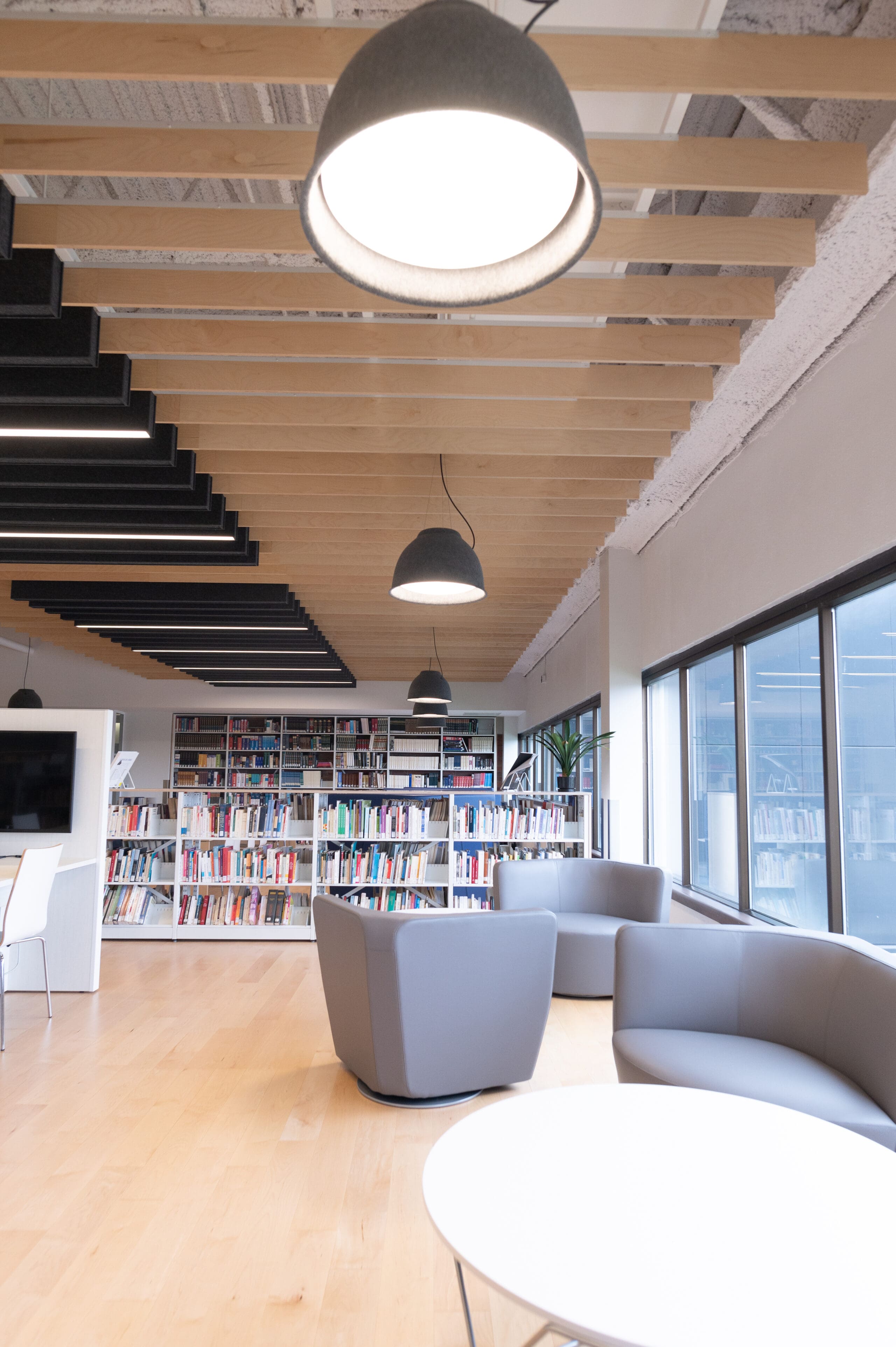 Un salon de bibliothèque moderne, avec des fauteuils gris, des tables rondes blanches, de grandes fenêtres, du parquet, des bibliothèques et des plafonniers, créant un espace lumineux et accueillant.