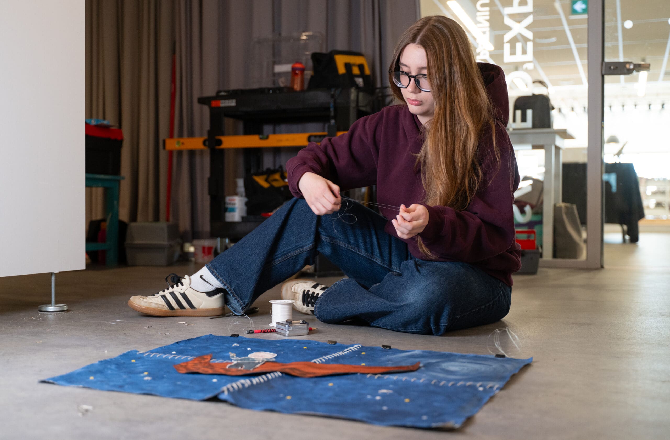 Une personne aux cheveux longs et portant des lunettes est assise par terre, en train de coudre du tissu avec une aiguille et du fil. Des ciseaux, du fil et des accessoires de couture sont posés sur le tissu bleu devant elle. Elle porte un jean, un sweat à capuche bordeaux et des baskets.