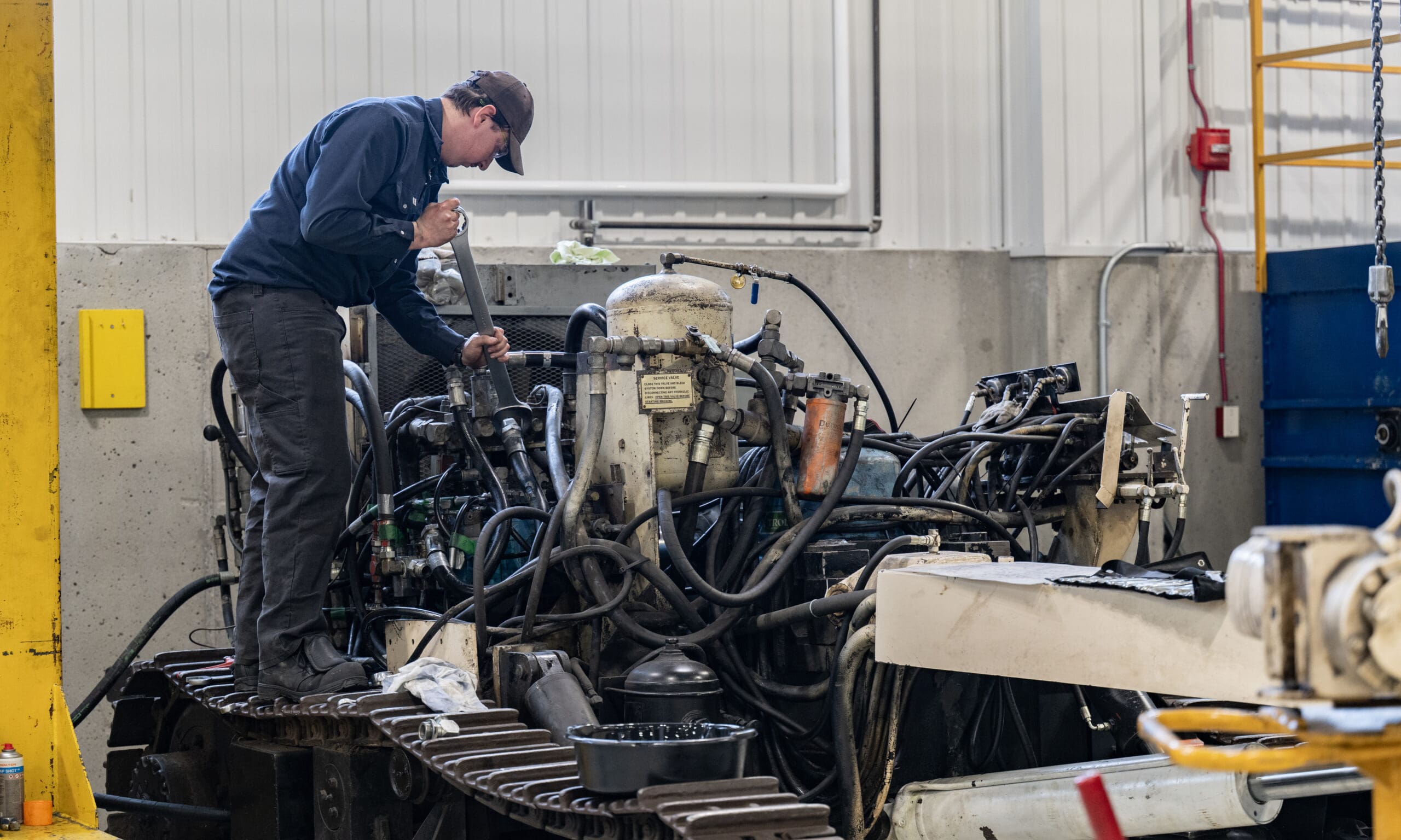 Une personne en tenue de travail se tient debout sur une machine, en train d'inspecter ou de réparer un gros moteur complexe, comportant de nombreux tuyaux et câbles, à l'intérieur d'un atelier industriel.
