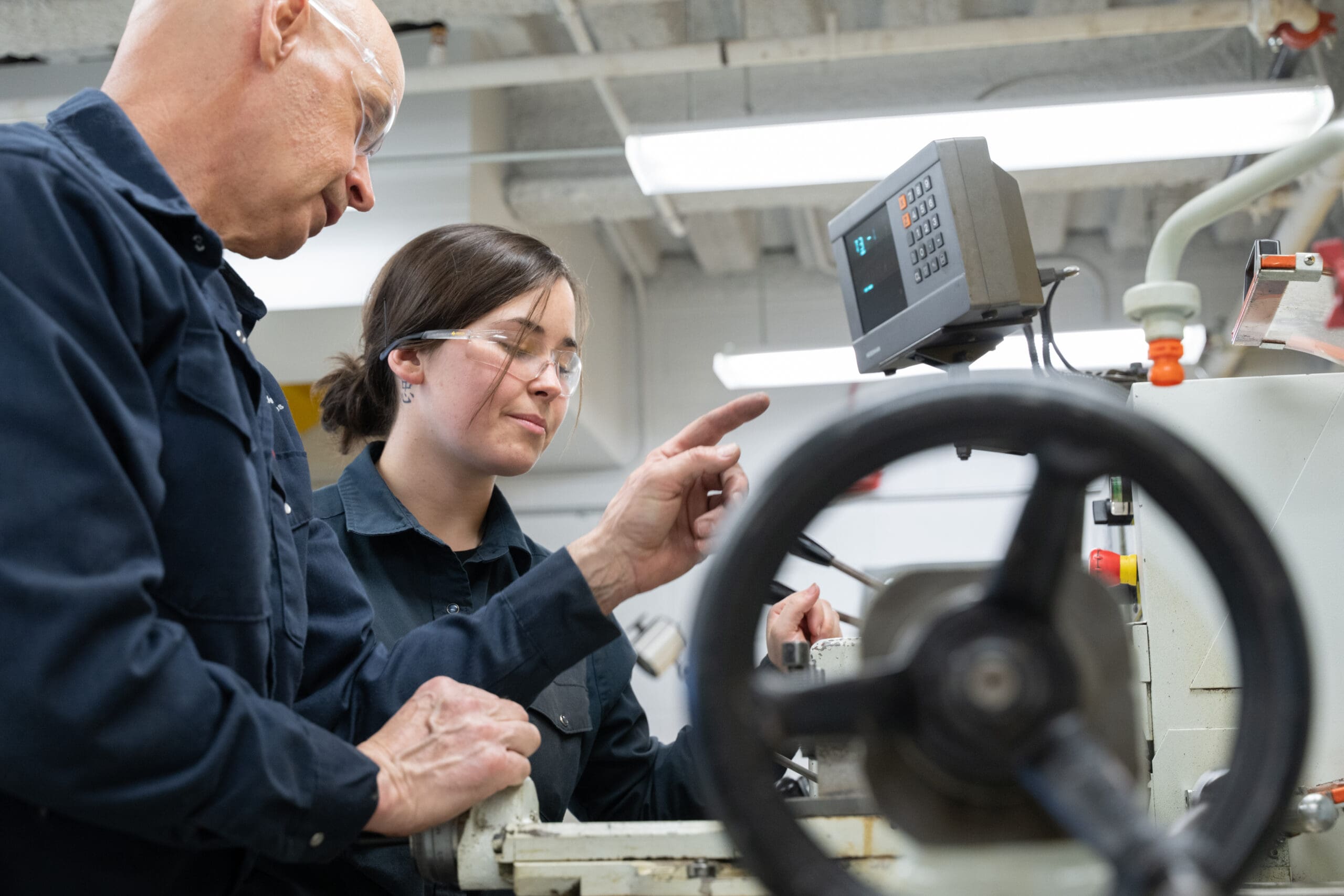 Deux personnes portant des lunettes de sécurité utilisent des machines dans un environnement industriel. L'une d'elles désigne un écran de commande tandis que l'autre observe ; toutes deux portent des uniformes de travail de couleur sombre.