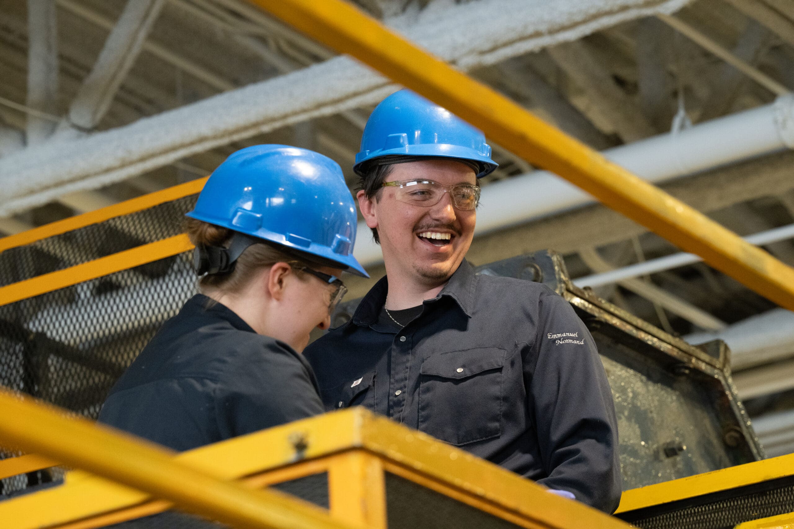 Deux personnes portant des casques de chantier bleus et des lunettes de sécurité sourient et discutent sur une plate-forme industrielle équipée de garde-corps jaunes.