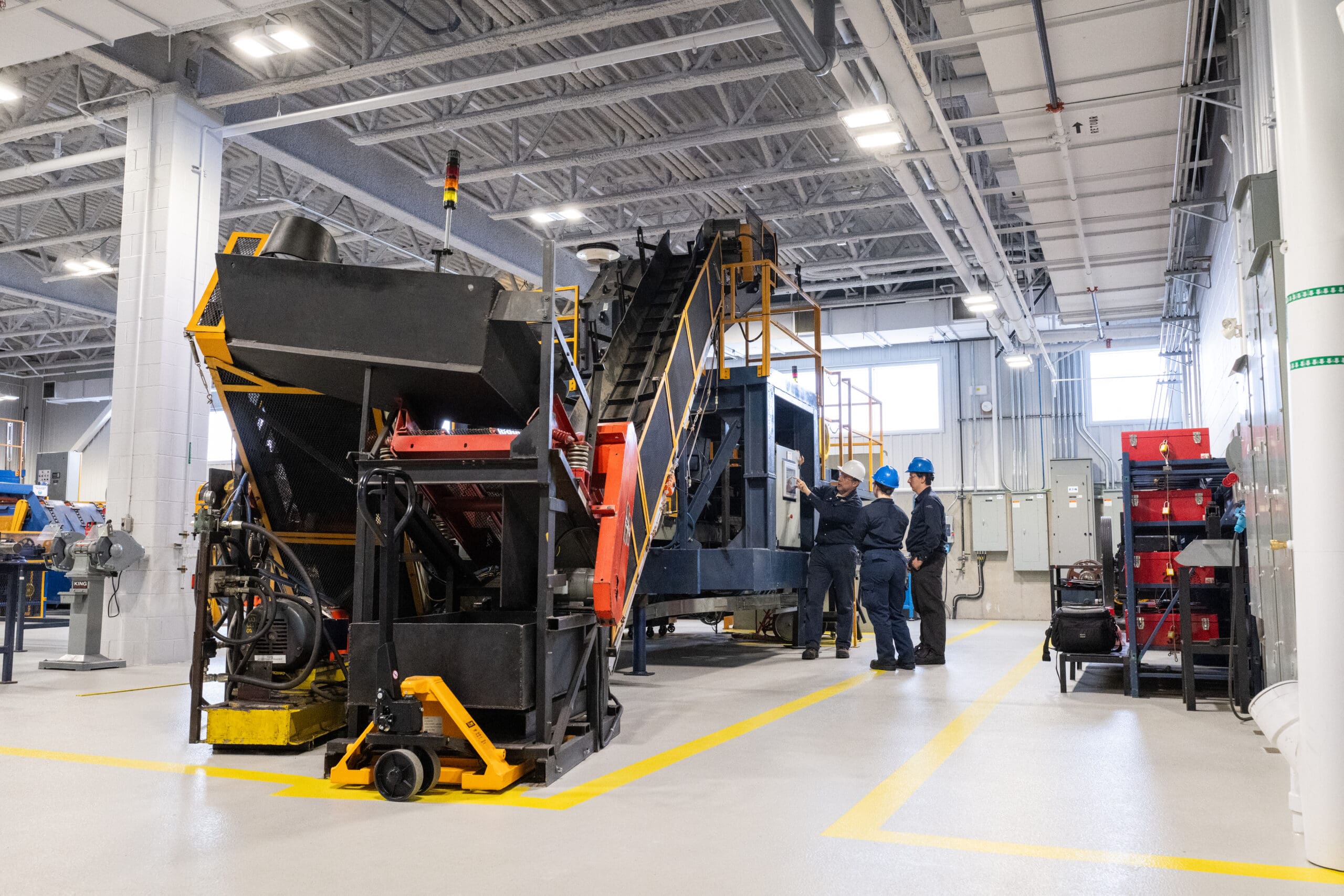 Trois personnes vêtues d'équipements de sécurité se tiennent à côté de grosses machines industrielles dans une usine lumineuse et spacieuse, dotée de hauts plafonds, de tuyaux métalliques et de divers équipements. Des lignes jaunes délimitent les allées sur le sol propre et de couleur claire.