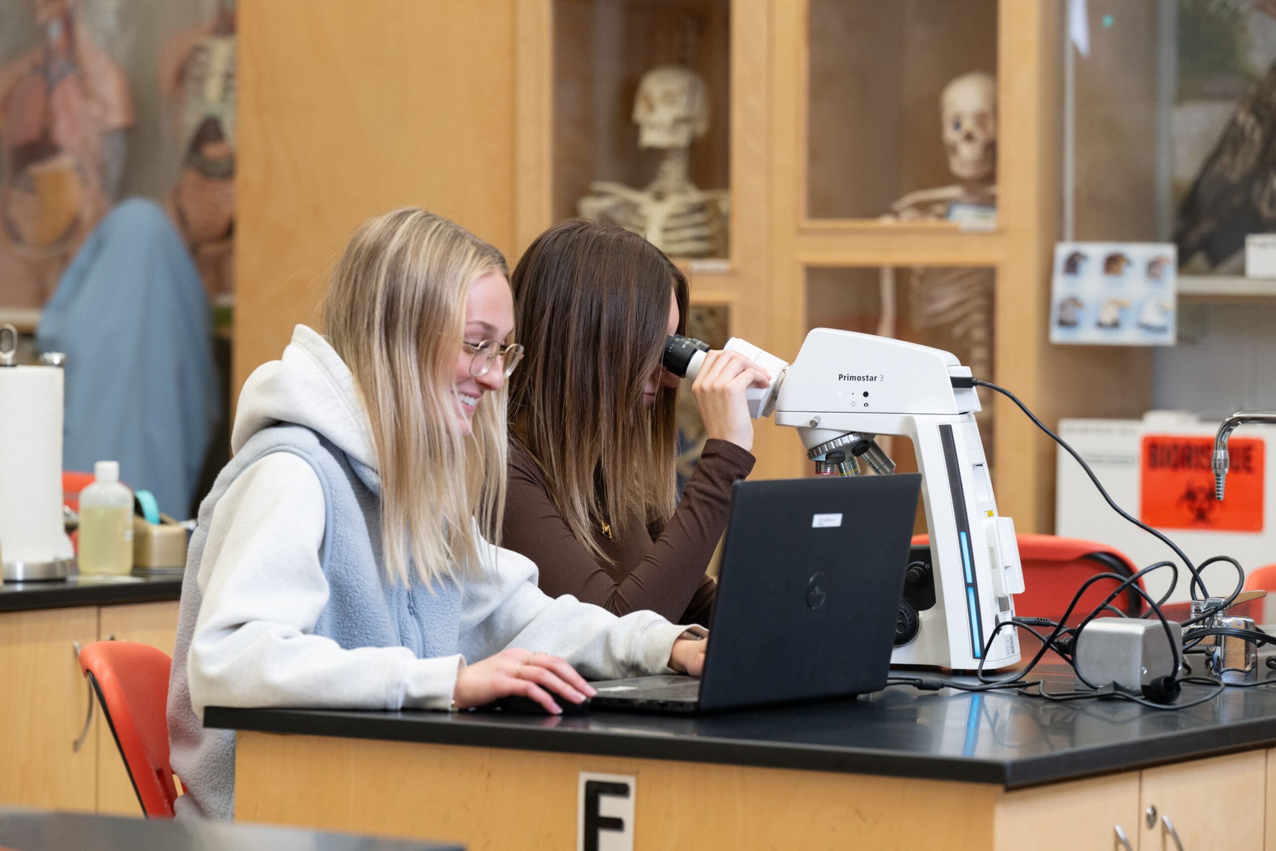 Deux élèves travaillent ensemble dans un laboratoire de sciences ; l'un utilise un ordinateur portable tandis que l'autre observe au microscope. On aperçoit des maquettes de squelettes et du matériel de laboratoire à l'arrière-plan.