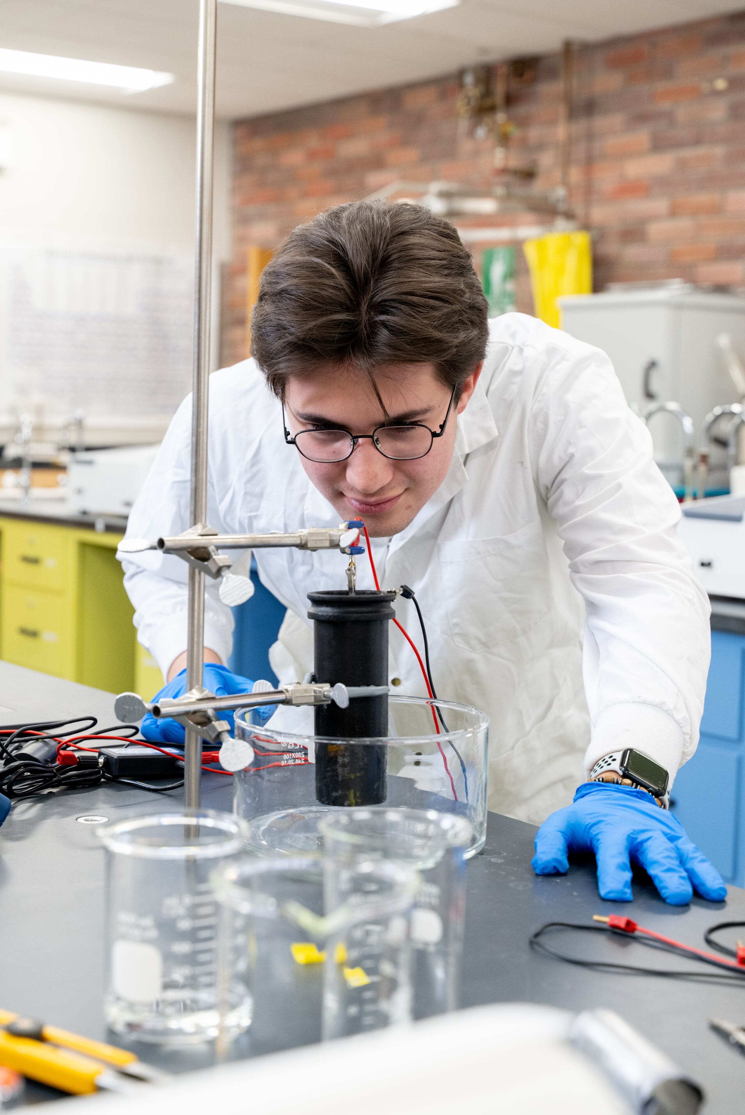 Une personne vêtue d'une blouse de laboratoire et portant des gants bleus observe une expérience scientifique menée à l'aide de béchers et d'appareils électriques posés sur une paillasse.