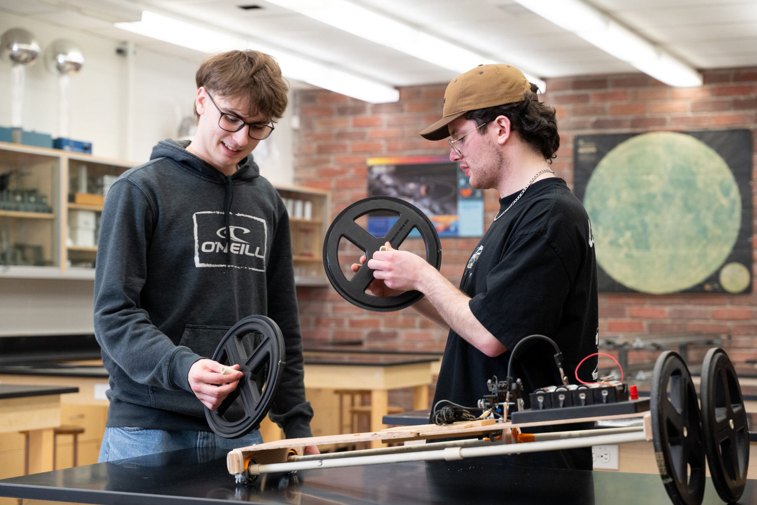 Dans une salle de classe, deux élèves examinent et discutent de grandes roues noires fixées à un robot en bois posé sur une table. L'un porte un sweat à capuche et des lunettes, l'autre une casquette et des lunettes. On aperçoit du matériel scientifique à l'arrière-plan.
