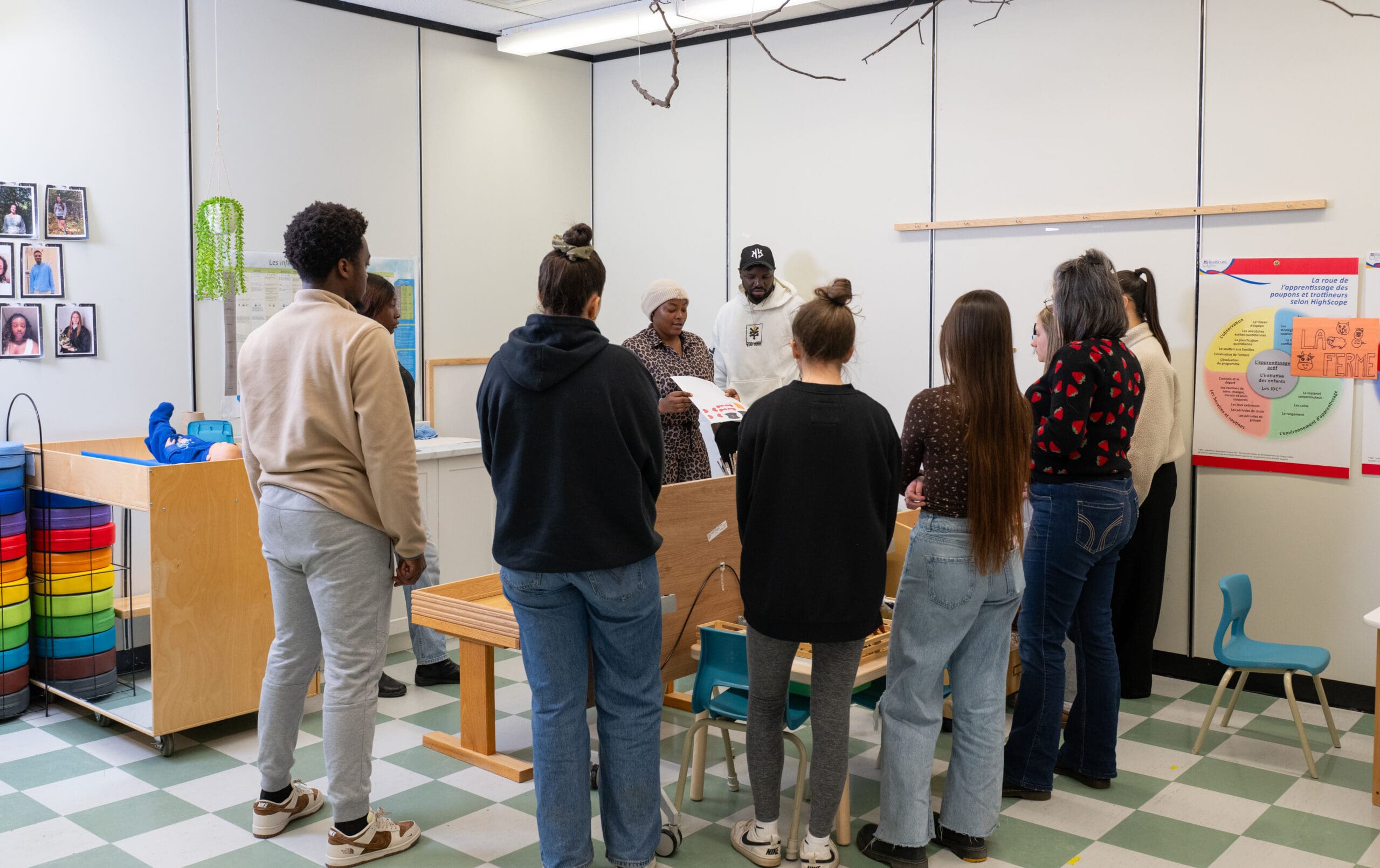 Un groupe de personnes se tient debout dans une salle de classe, écoutant une femme qui tient un livre. La pièce comporte des rangements colorés, des photos accrochées au mur et des affiches pédagogiques. Certaines personnes tournent le dos à la caméra.
