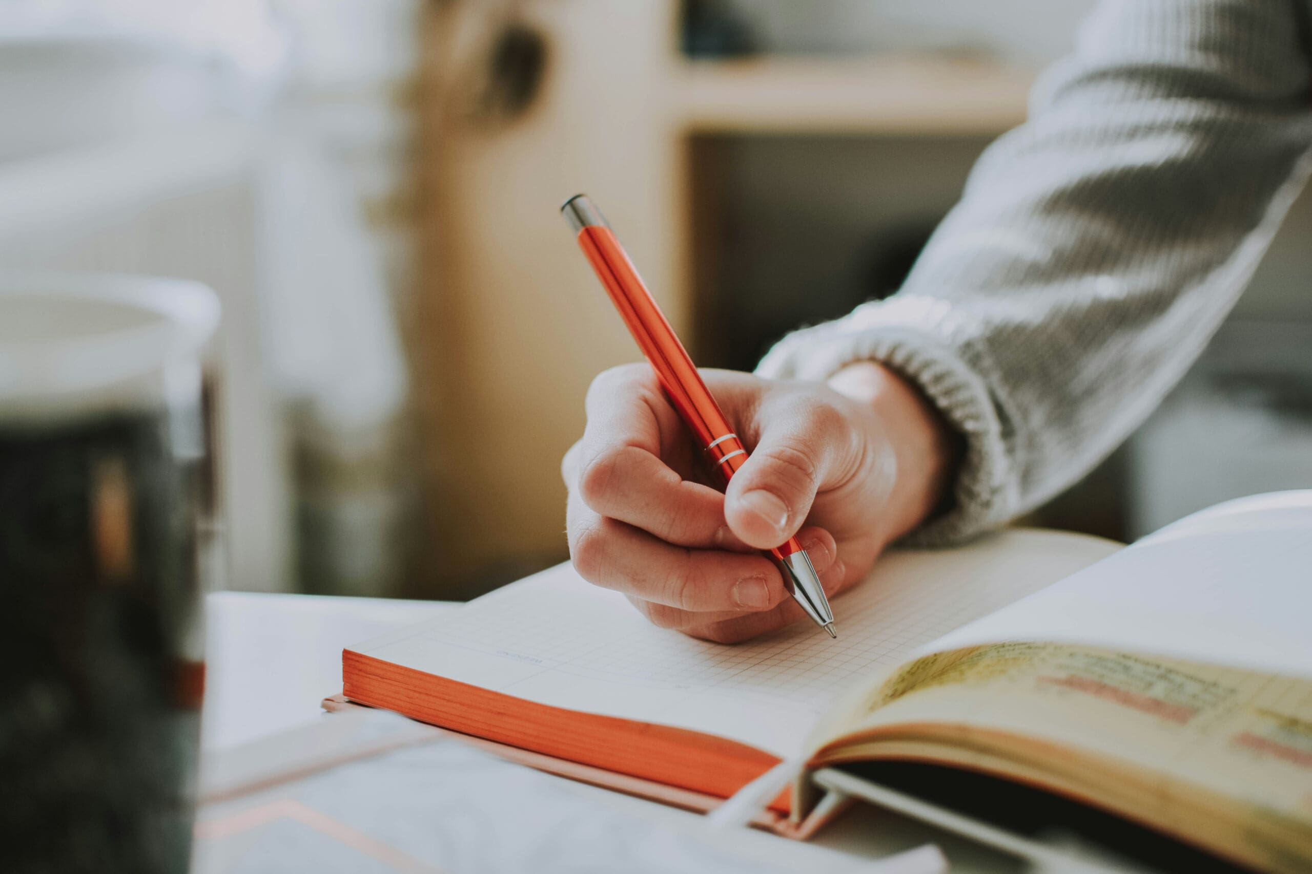 Une personne vêtue d'un pull gris écrit au stylo rouge dans un cahier à quadrilles. Un livre ouvert et une partie d'une tasse sont posés sur le bureau à proximité.