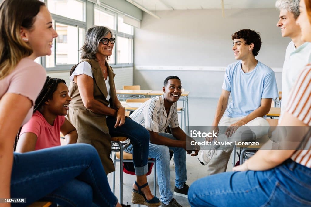 Un enseignant et cinq élèves sont assis dans une salle de classe ; ils sourient et discutent ensemble dans une ambiance détendue et conviviale. On aperçoit des pupitres et de grandes fenêtres à l'arrière-plan.