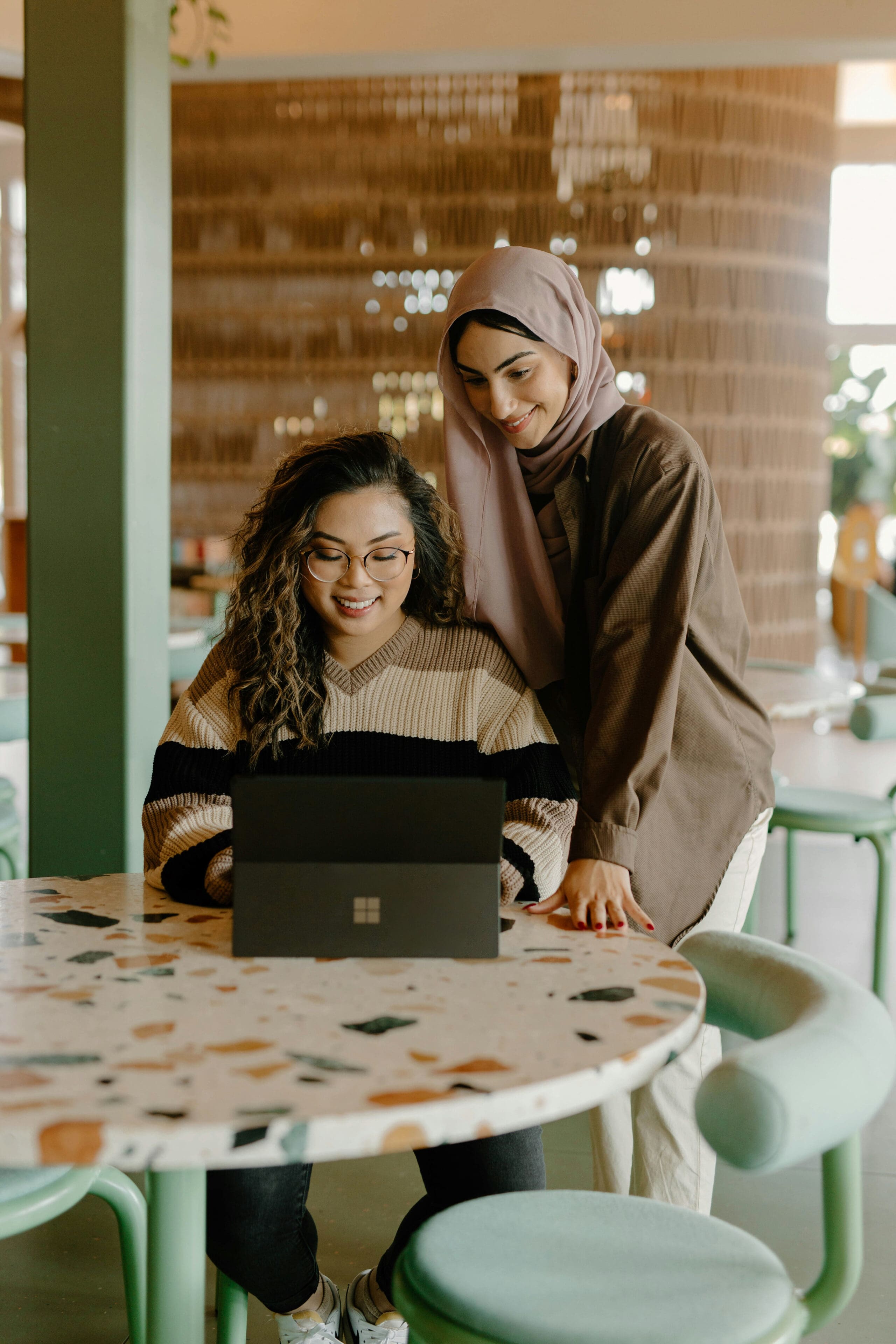 Deux femmes souriantes regardent ensemble un ordinateur portable dans un café moderne et bien éclairé. L'une est assise et porte des lunettes, tandis que l'autre se tient debout à côté d'elle et porte un hijab.