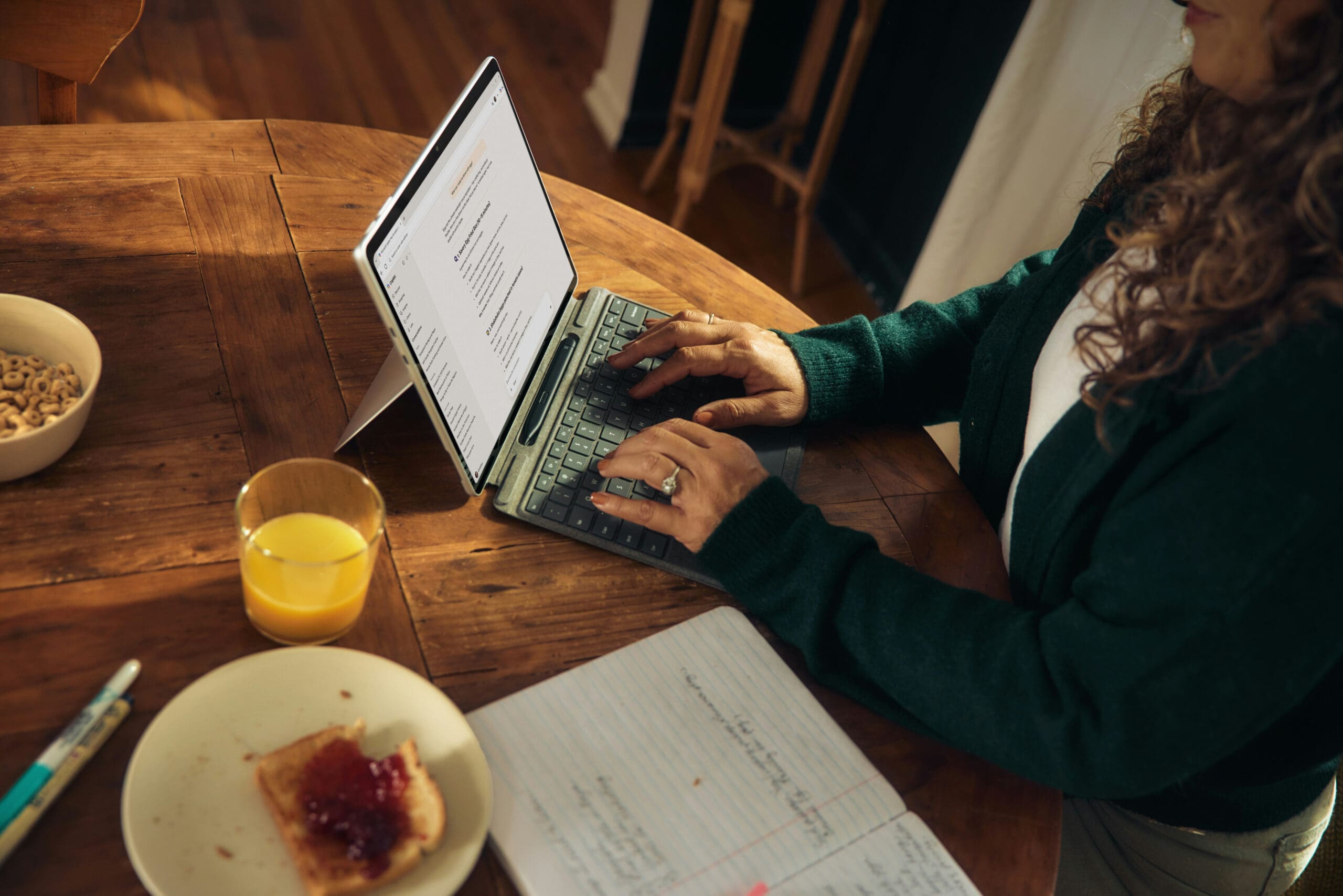 Une personne tapant sur un ordinateur portable, assise à une table en bois, avec à côté d'elle un cahier, un stylo, une tartine à la confiture, un bol de céréales et un verre de jus d'orange. Seules ses mains et une partie de son torse sont visibles.
