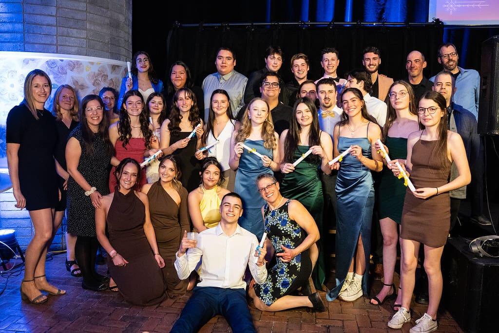 Un groupe de personnes, principalement des jeunes adultes, posent ensemble à l'intérieur, en tenue de cérémonie ; certaines tiennent des documents roulés ou des certificats. Elles sourient dans une ambiance de fête, avec des rideaux sombres et un éclairage tamisé.
