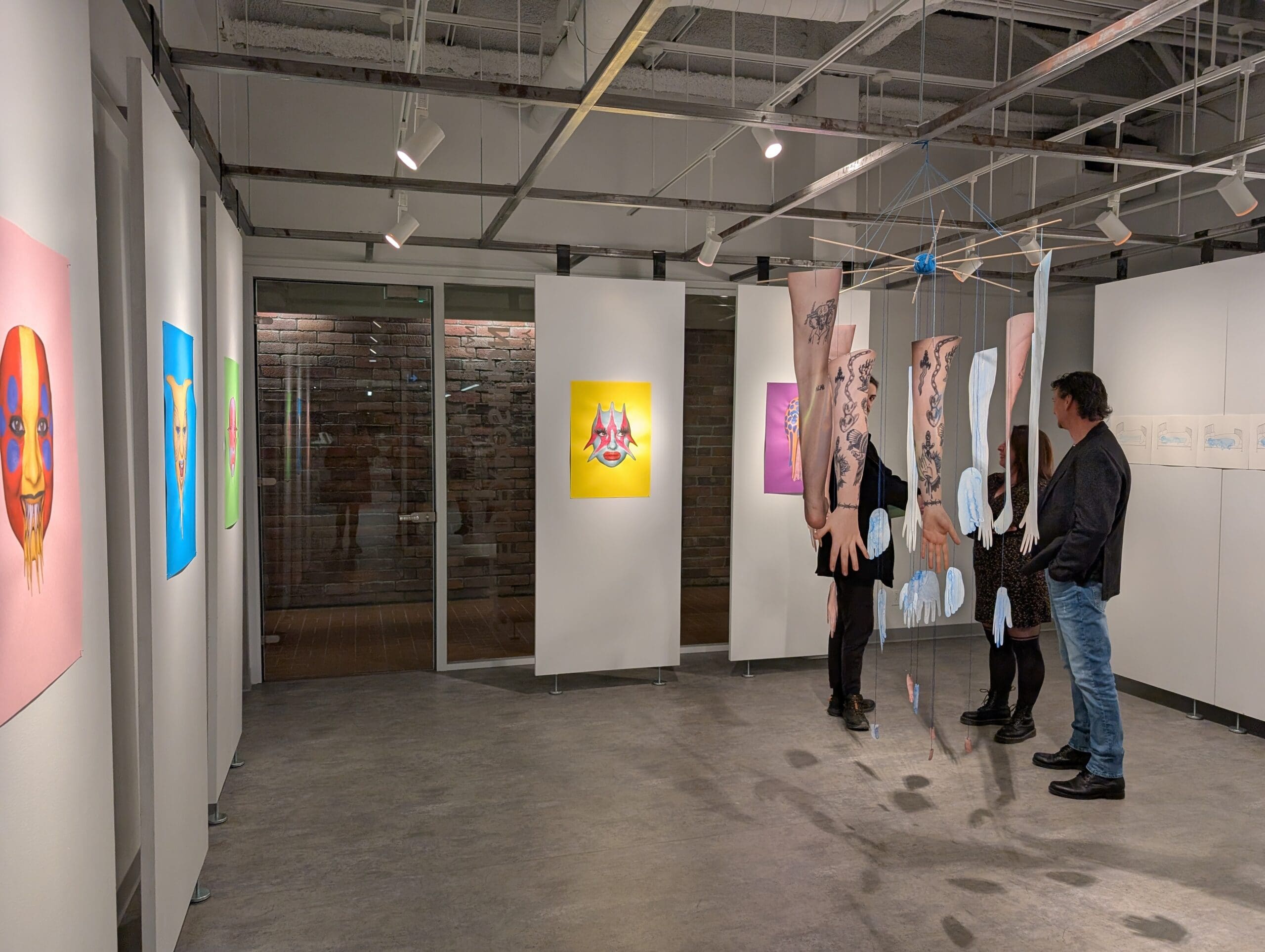 Des visiteurs admirent des œuvres d'art colorées et surréalistes accrochées aux murs blancs de la galerie. Trois personnes se tiennent près d'œuvres suspendues représentant des membres humains. Des lumières vives éclairent cet espace artistique moderne, de style industriel.