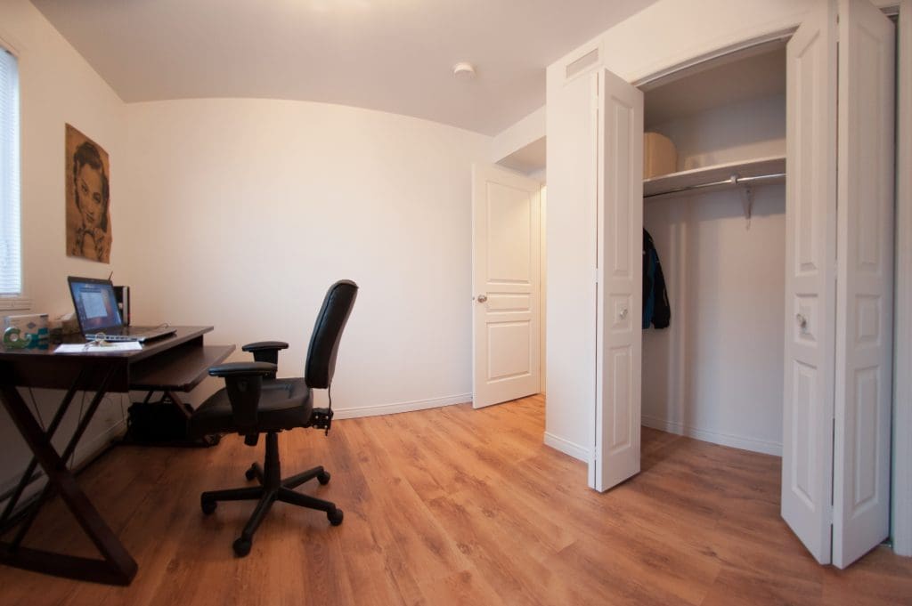 Une chambre bien rangée avec du parquet, un bureau et une chaise à gauche, une petite armoire ouverte où est posée une veste à droite, et un portrait accroché au mur. La lumière naturelle pénètre par une fenêtre située près du bureau.