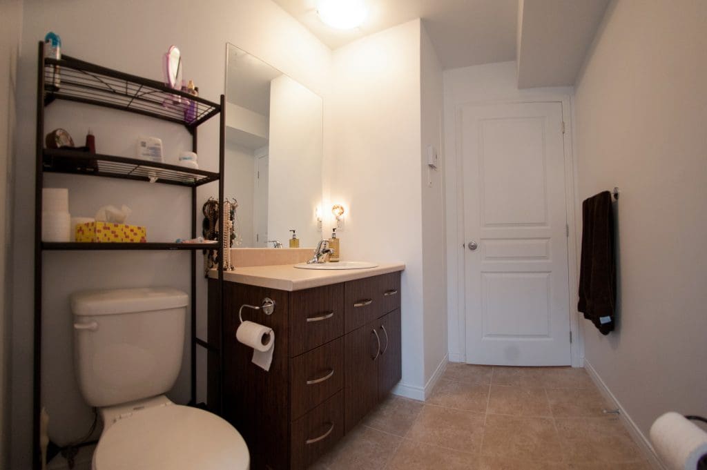 Salle de bains moderne équipée de toilettes, d'une étagère métallique, d'un meuble-lavabo marron avec un lavabo, d'un miroir et d'un plafonnier. Une serviette marron est accrochée au mur et le sol est carrelé de beige.