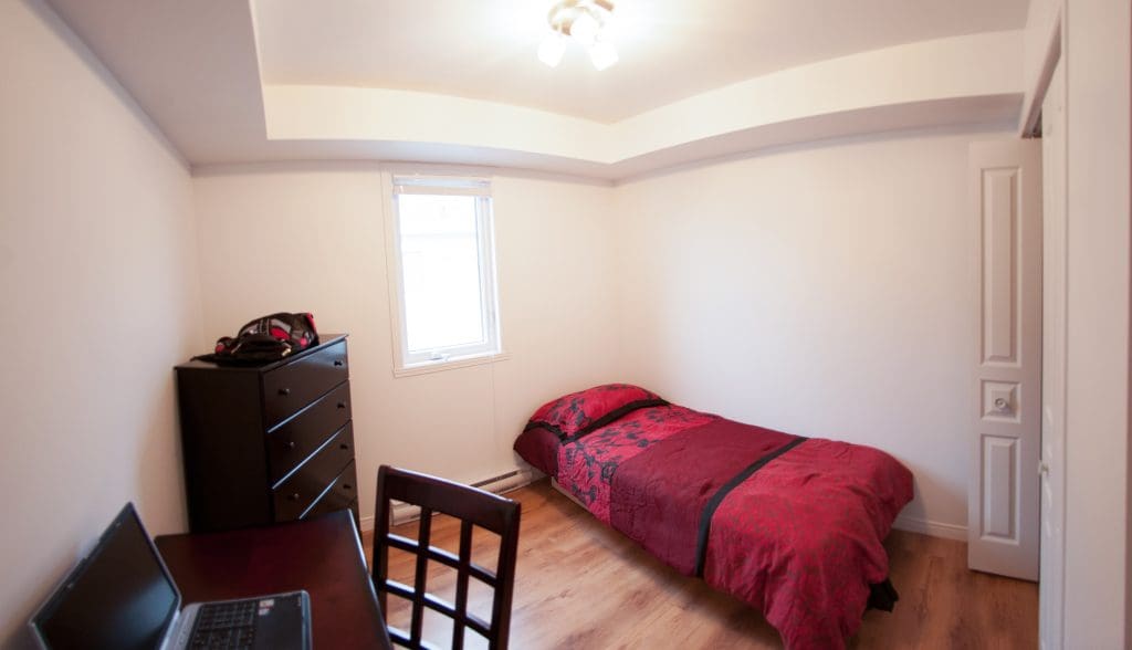 Une petite chambre avec un lit simple recouvert d'une parure de lit rouge, une commode foncée, un ordinateur portable posé sur un bureau, une chaise, un parquet et une fenêtre laissant entrer la lumière naturelle.