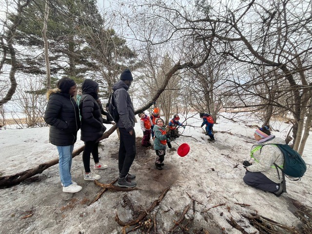 Un groupe d'adultes et d'enfants se rassemble dans une zone boisée enneigée ; l'un des enfants tient un disque en plastique rouge.
