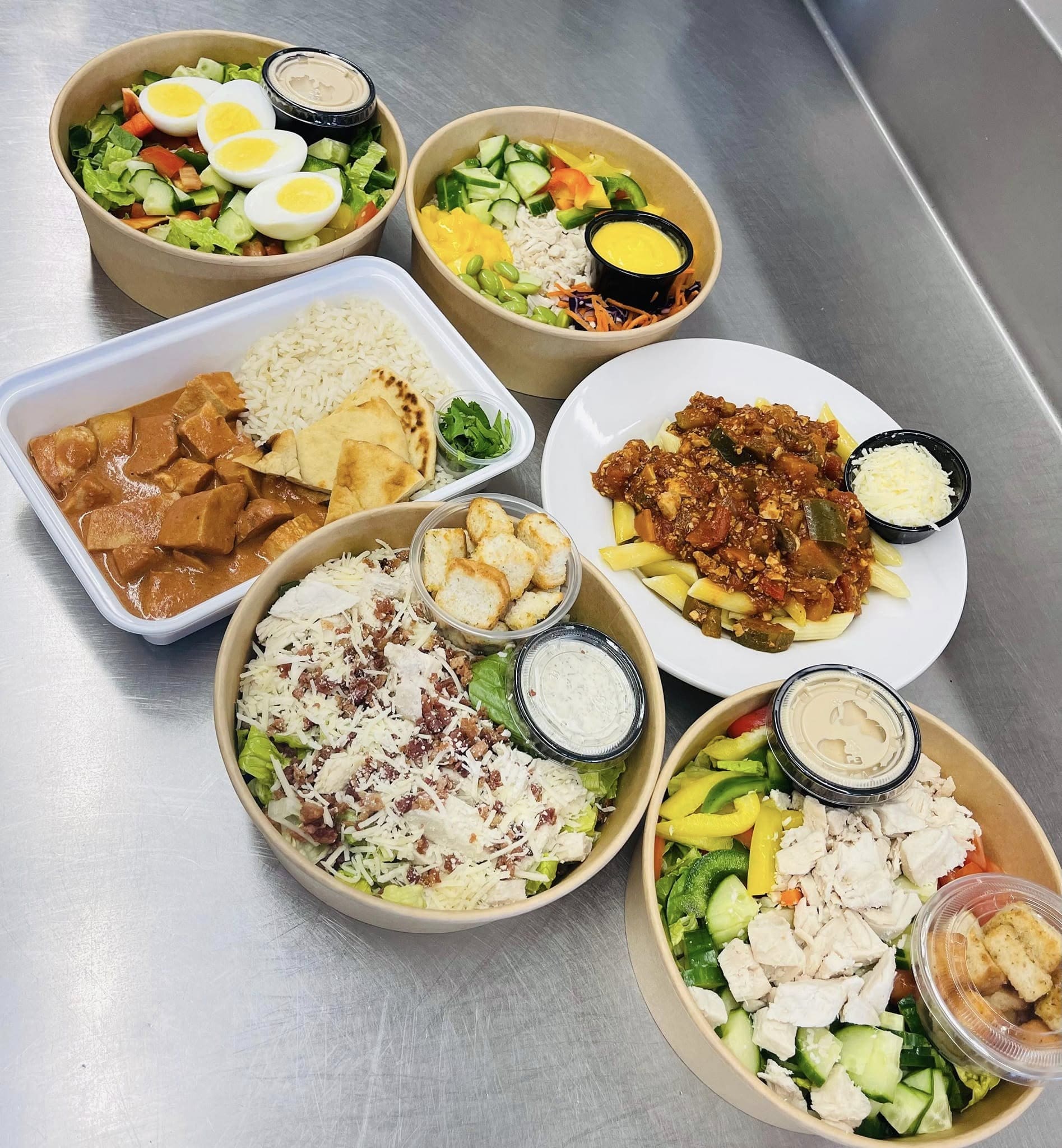 Cinq salades et deux plats chauds sont disposés sur un plateau en métal. Les salades sont composées d'œufs, de croûtons, de dinde, de fromage et de petits pots de vinaigrette. Les plats chauds comprennent du riz à la sauce brune et un plat de viande accompagné de légumes.