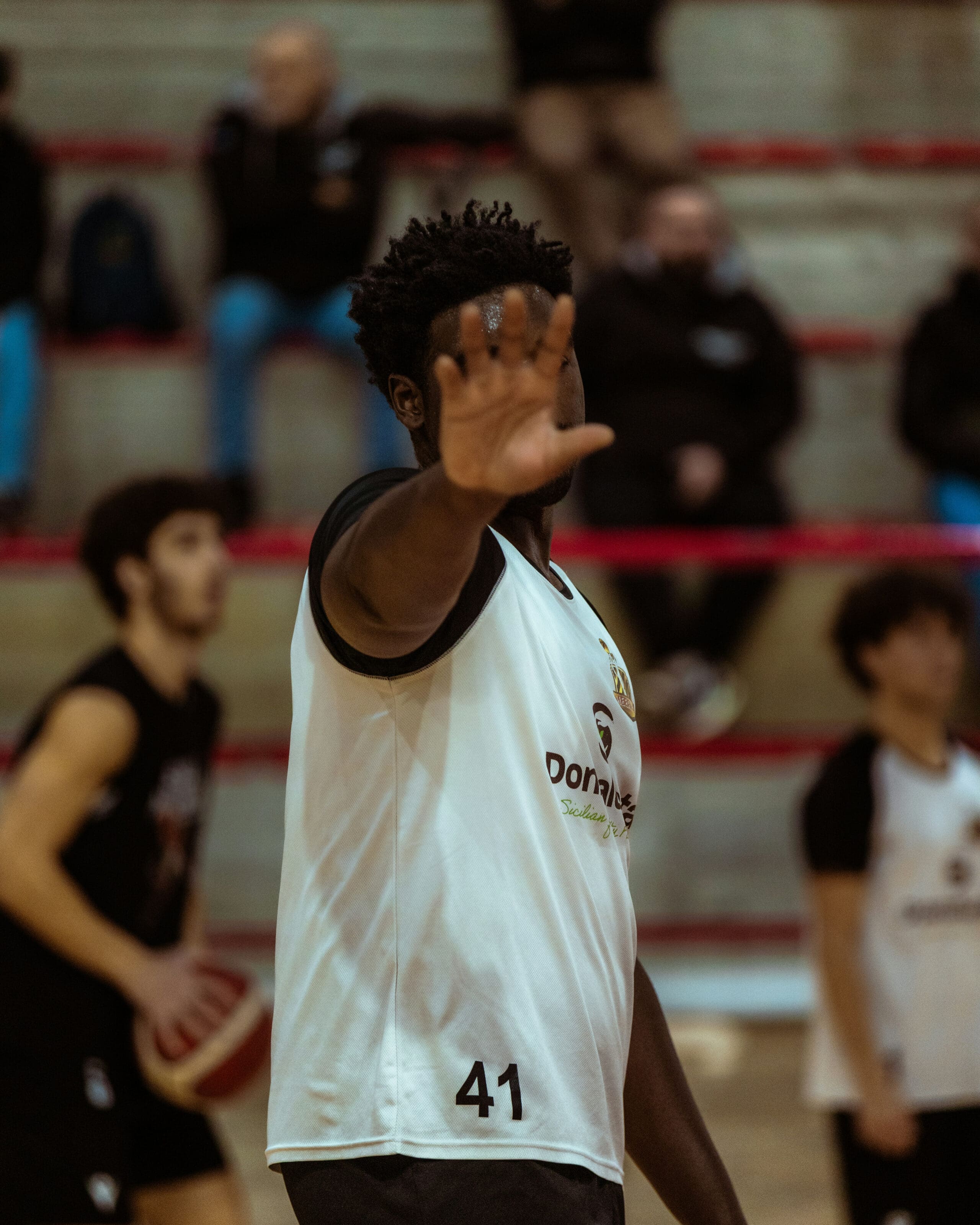 Un joueur de basket-ball vêtu d'un maillot blanc portant le numéro 41 se tient sur le terrain, la main levée pour faire écran à l'objectif. D'autres joueurs et des spectateurs sont visibles à l'arrière-plan, flous.