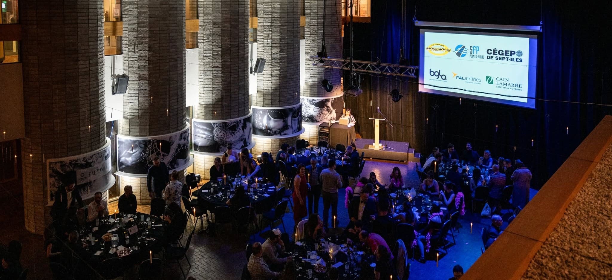 Un événement officiel où les invités sont assis à des tables rondes, avec un intervenant à la tribune et un grand écran affichant les logos des sponsors, dans une salle spacieuse et faiblement éclairée.
