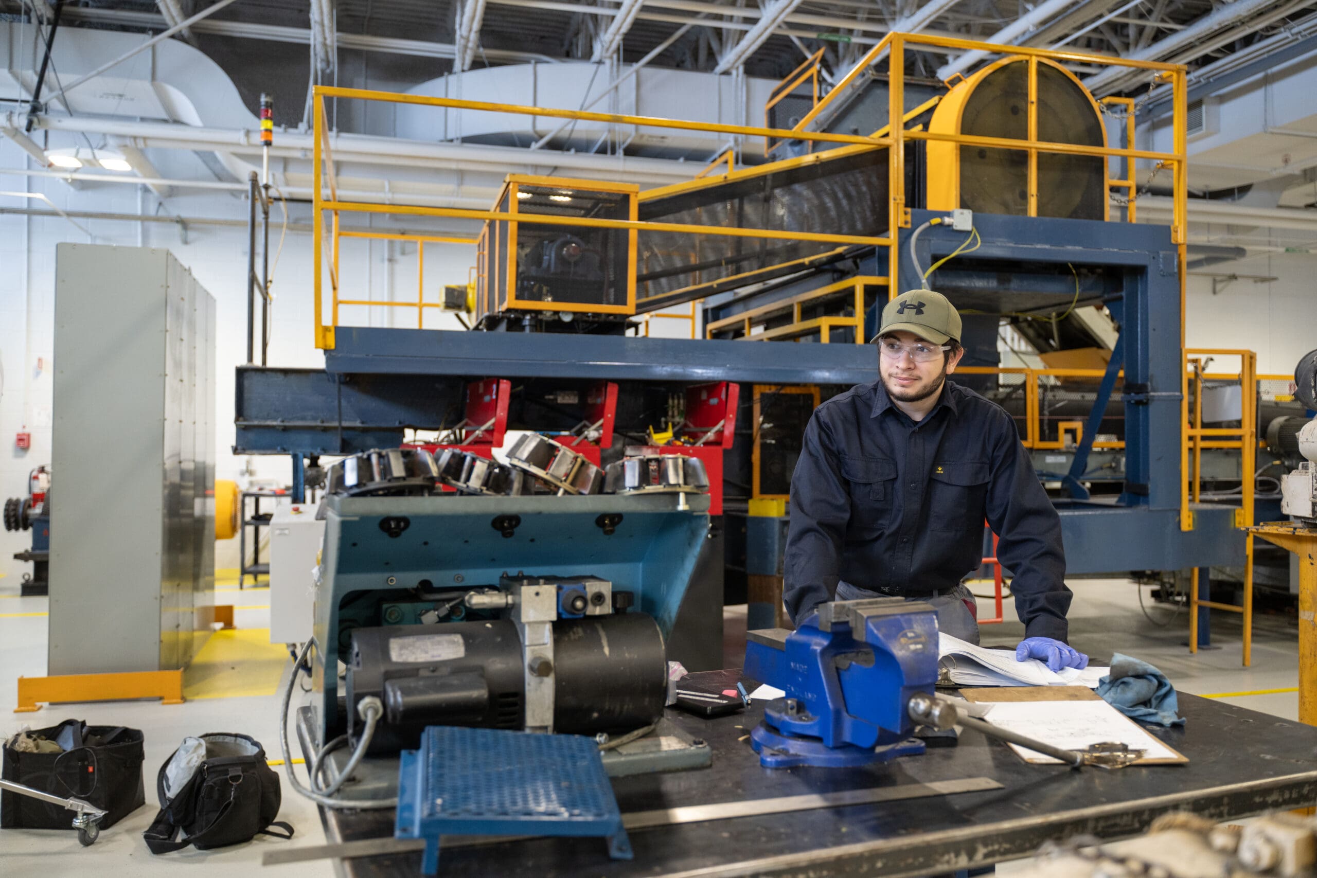 Une personne vêtue d'une tenue de travail et portant des gants se tient debout devant un établi métallique dans un site industriel, entourée de machines et d'équipements. L'espace est très éclairé et on aperçoit des structures suspendues au plafond.