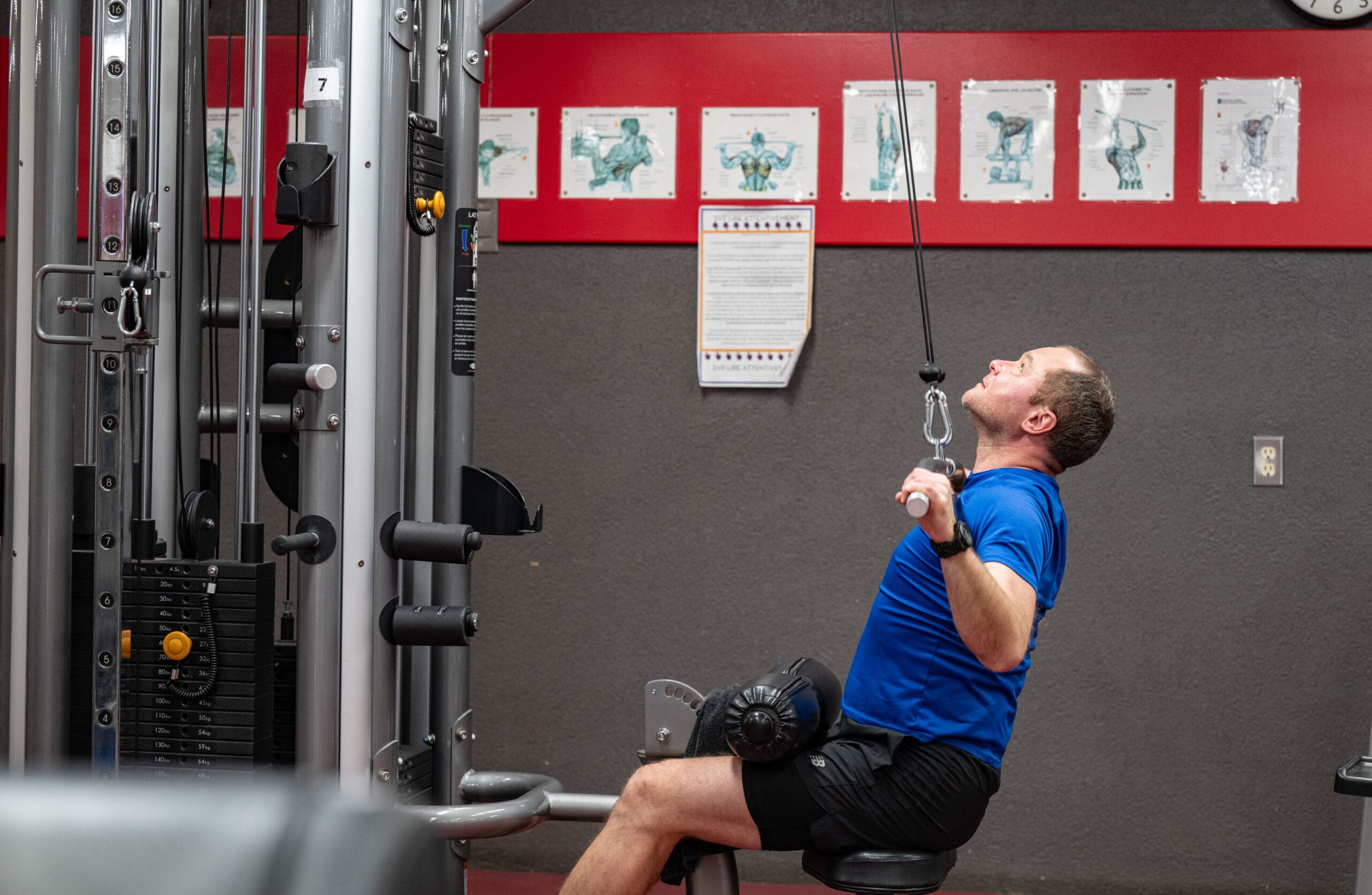 Un homme en chemise bleue est assis sur un appareil de musculation et tire sur un câble pour effectuer un exercice de tirage dorsal. Des schémas d'entraînement sont visibles sur le mur derrière lui.