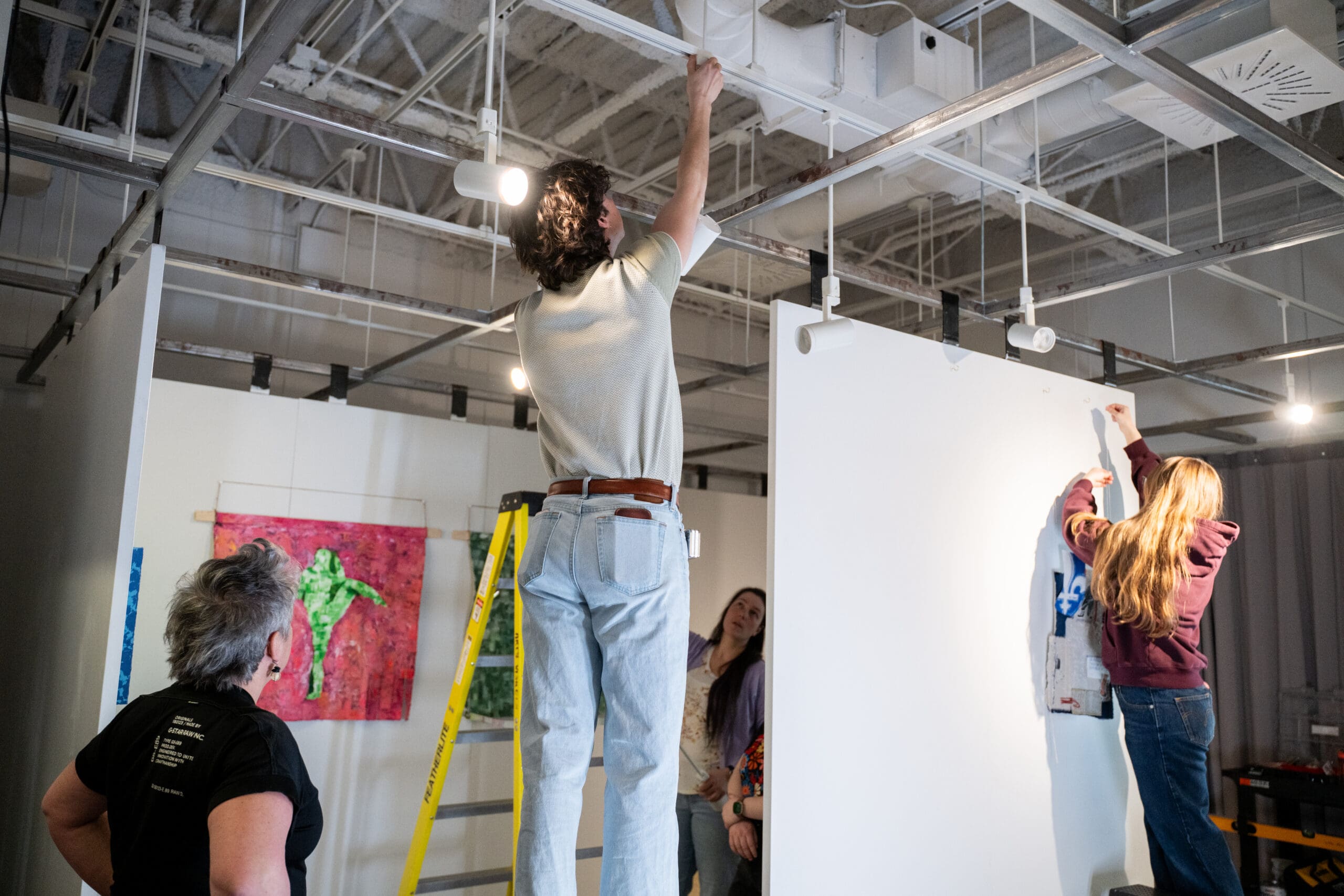 Quatre personnes sont en train d'aménager une galerie d'art ; deux d'entre elles sont perchées sur des échelles, occupées à régler l'éclairage et à accrocher des œuvres, tandis que les deux autres les observent. Des œuvres d'art aux couleurs vives ornent les murs. Le plafond est parcouru de tuyaux et de luminaires apparents.