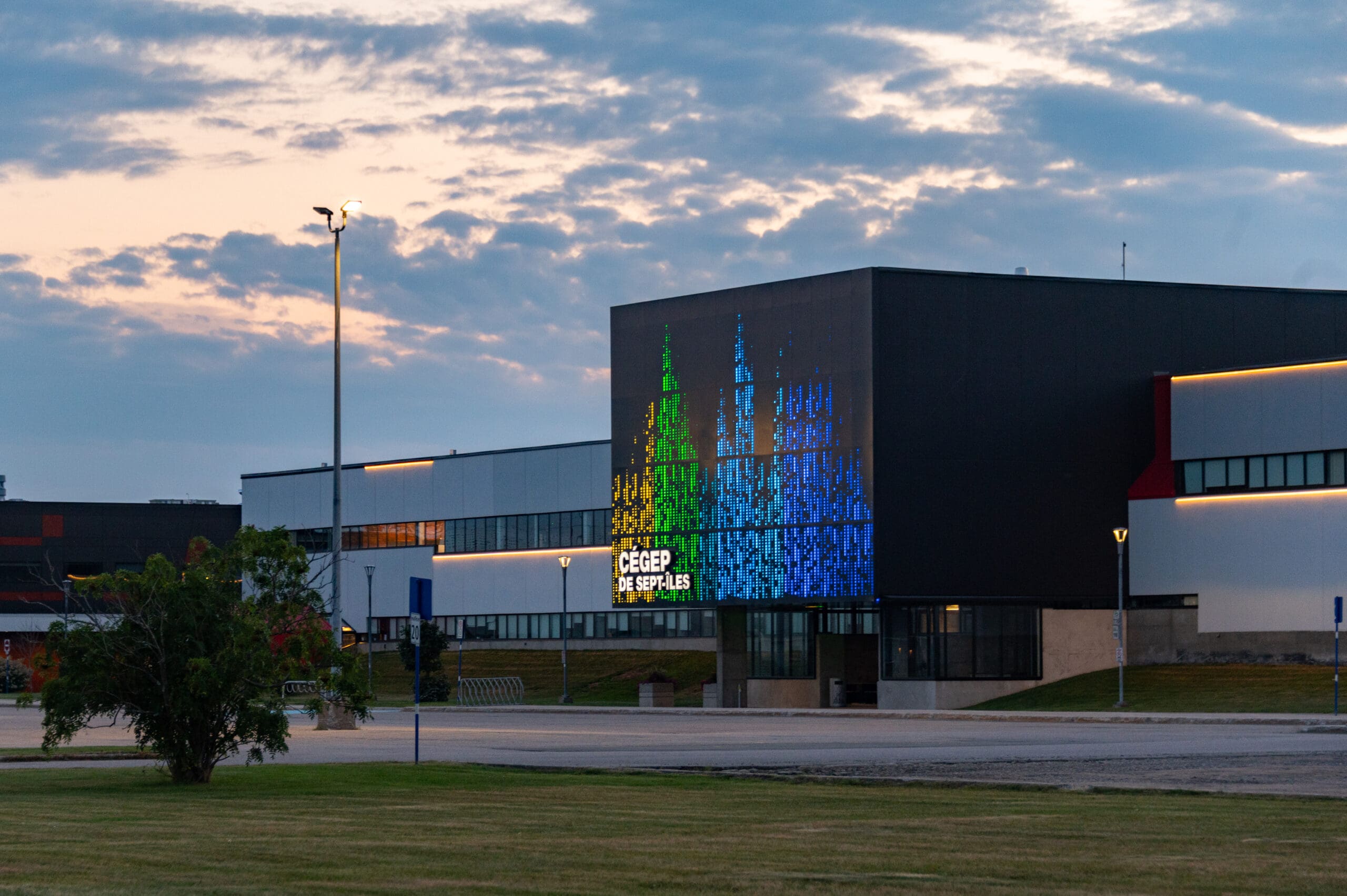 Un bâtiment moderne sur lequel, à la tombée de la nuit, un grand écran numérique affiche des lignes verticales colorées et l'inscription « Cégep de Sept-Îles », avec un ciel nuageux et une pelouse au premier plan.