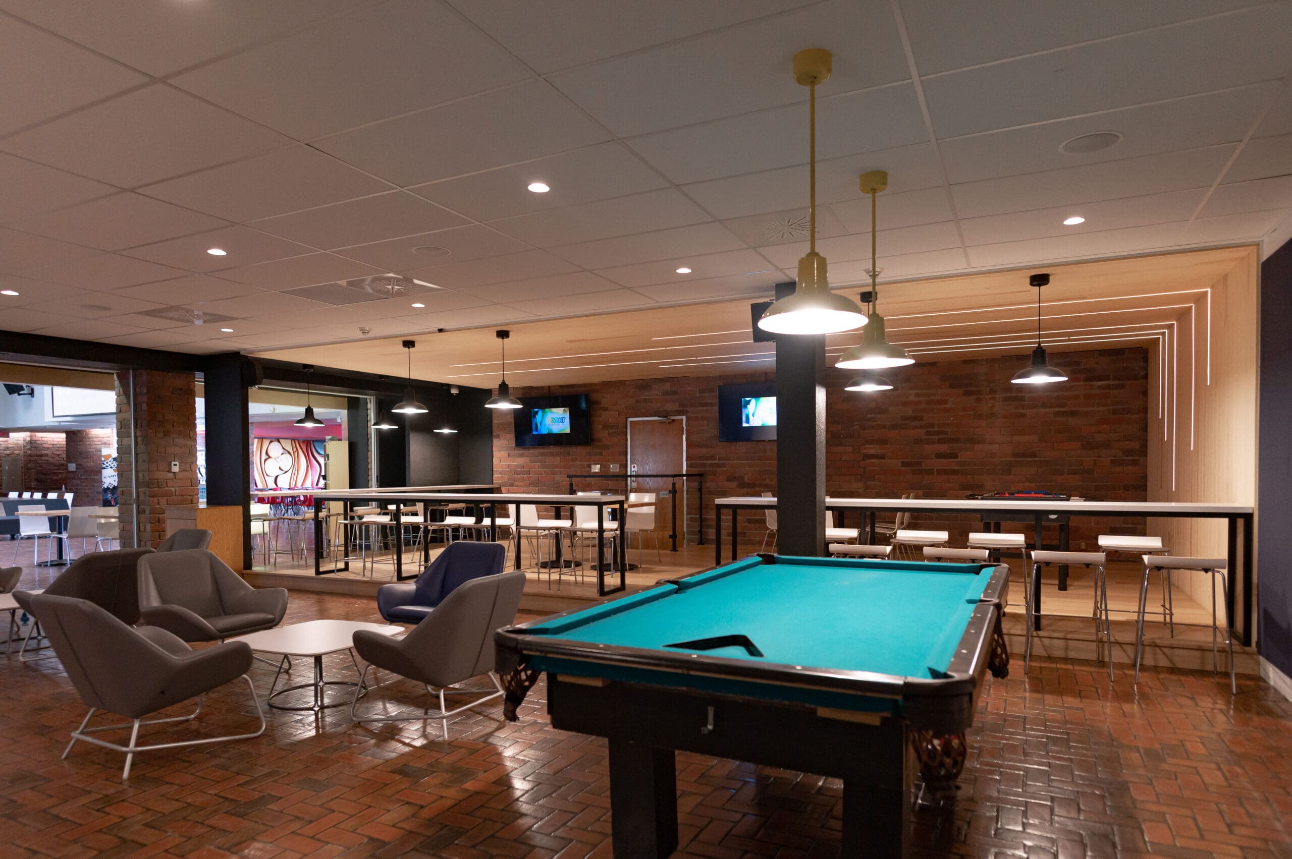 Un salon moderne avec un billard au premier plan, entouré de chaises grises, de tables, de tabourets hauts, de murs en briques et de suspensions. Deux téléviseurs sont fixés au mur du fond.
