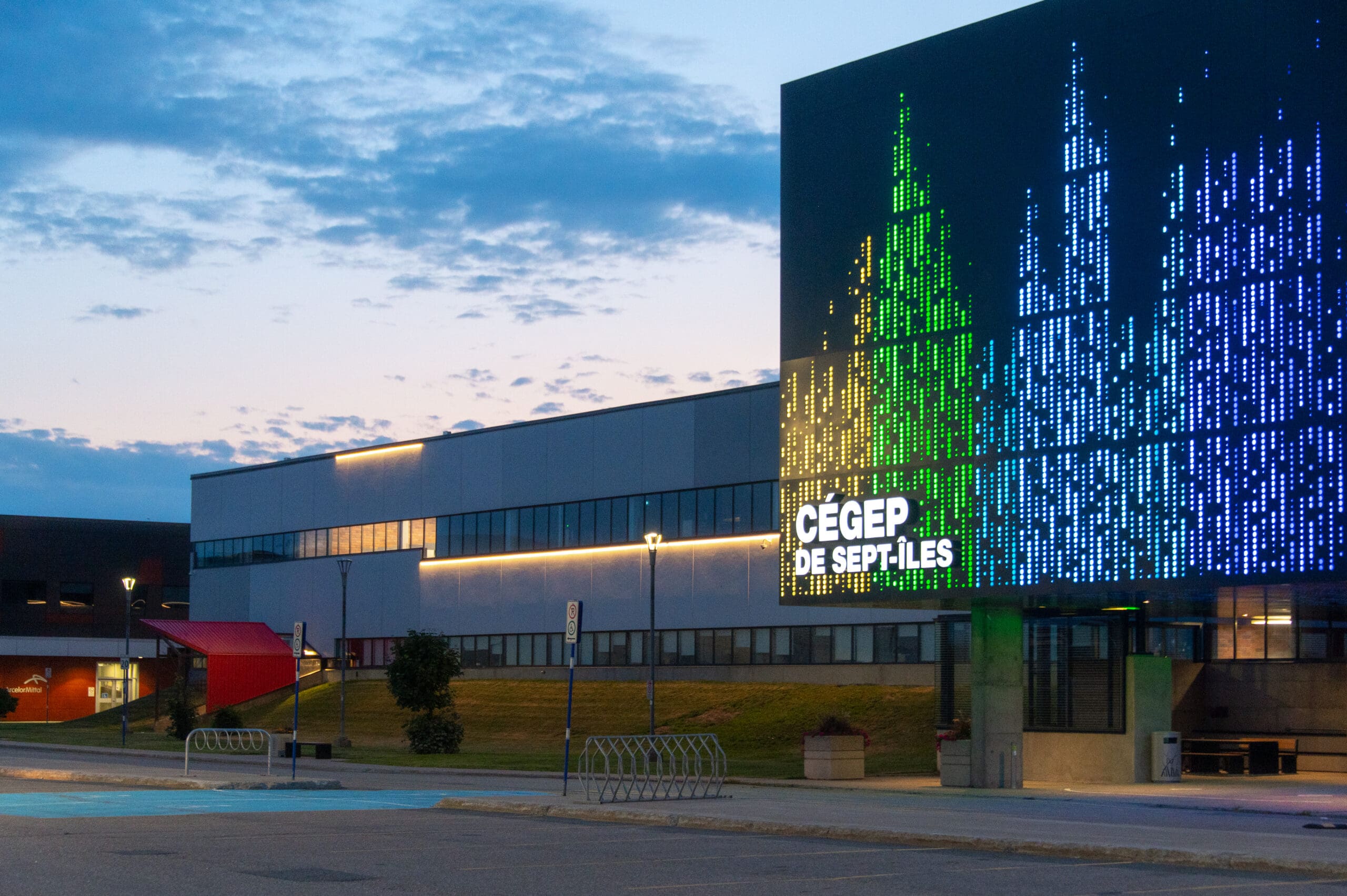 Le bâtiment du Cégep de Sept-Îles au crépuscule, avec un affichage numérique de lumières colorées sur la façade, un porte-vélos et un parking vide au premier plan.