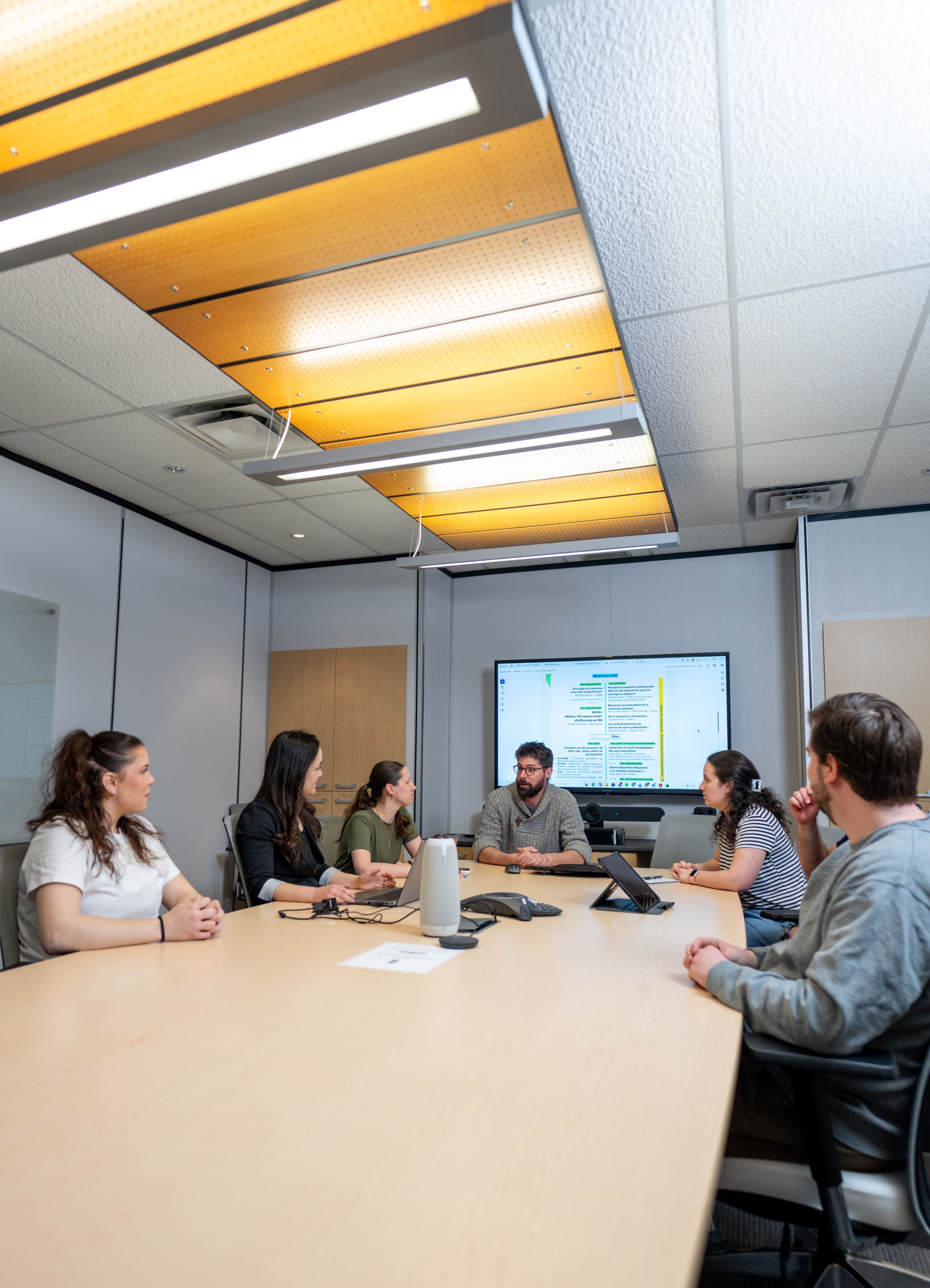 Six personnes sont assises autour d'une table de conférence dans une salle de réunion et discutent. Une présentation comprenant du texte et des graphiques s'affiche sur un grand écran situé au bout de la table. La pièce est éclairée par de puissants plafonniers.