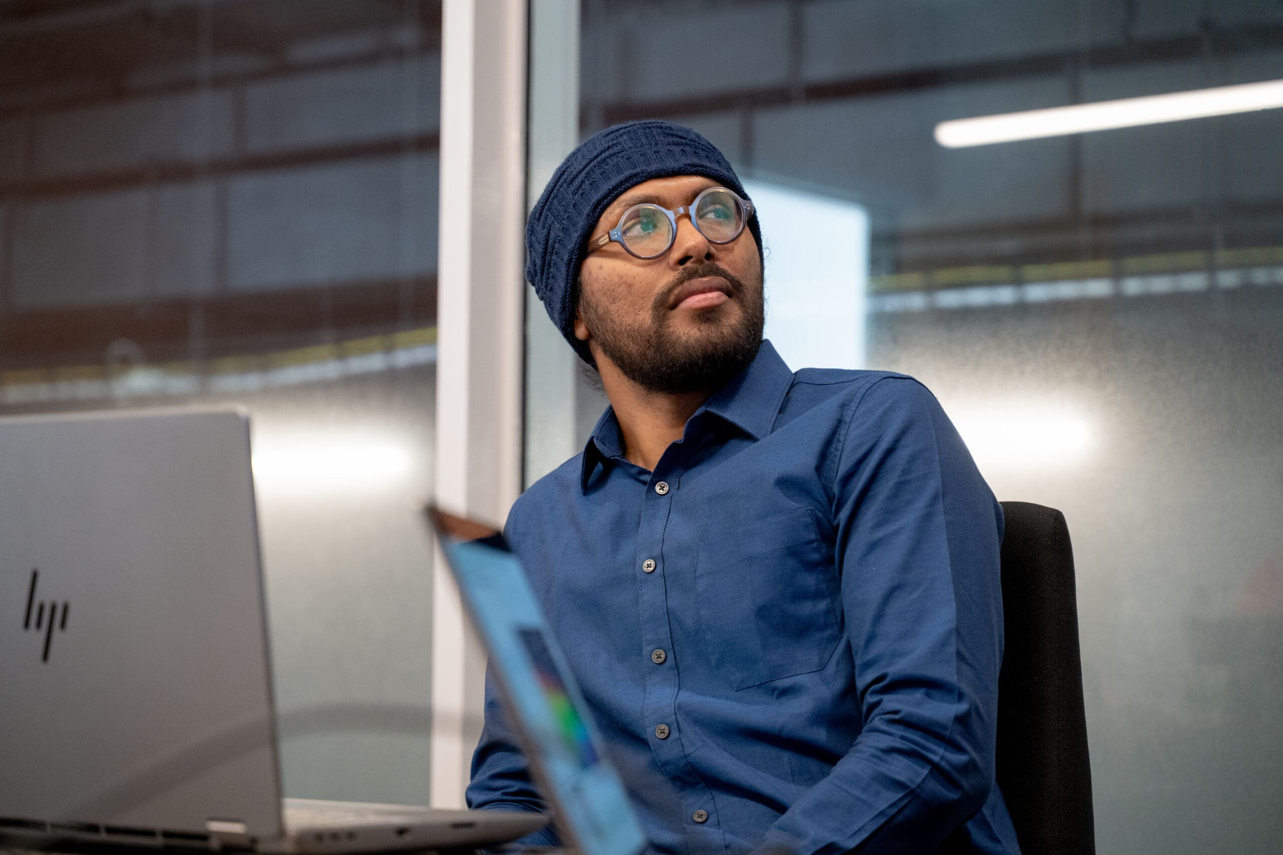 Un homme à la barbe foncée et portant des lunettes, vêtu d'une chemise bleue et d'un bonnet en tricot, est assis à un bureau devant un ordinateur portable et lève les yeux, dans un décor de bureau.