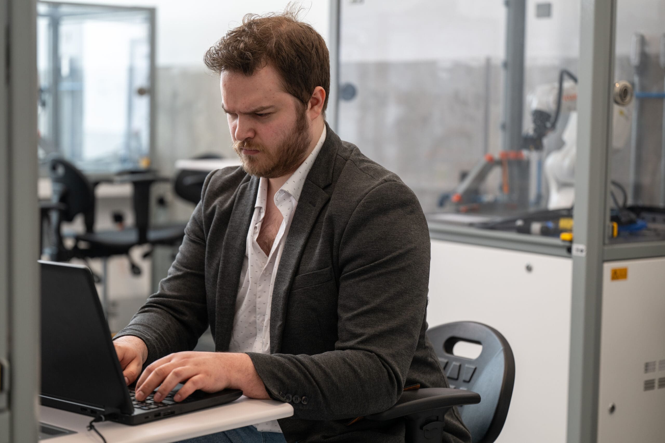 Un homme barbu, vêtu d'un blazer gris, travaille sur un ordinateur portable, assis à un bureau dans un bureau ou un laboratoire moderne, avec du matériel et des chaises visibles à l'arrière-plan.