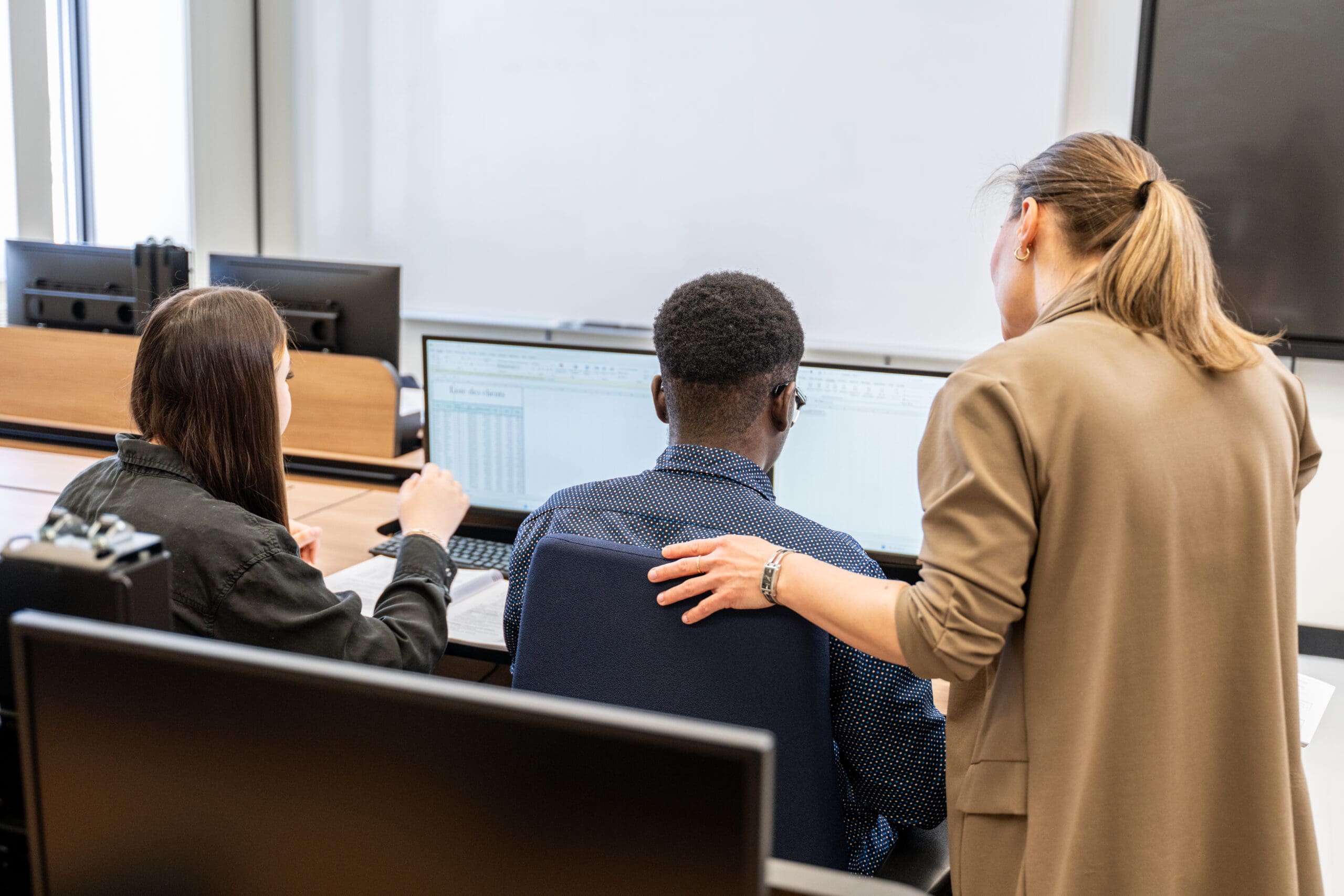 Dans une salle de classe, trois personnes sont assises devant un ordinateur. L'une d'elles se lève et pose délicatement la main sur l'épaule d'une autre, comme pour lui donner des conseils, tandis que tout le monde regarde l'écran de l'ordinateur.