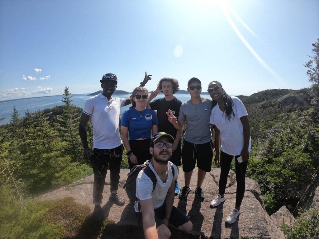 Six amis prennent la pose et sourient pour une photo de groupe sur une corniche rocheuse entourée d'arbres, avec en arrière-plan une vue panoramique sur les collines, la forêt et l'eau, par une journée ensoleillée.