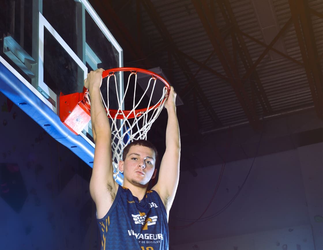 Un joueur de basket-ball vêtu d'un maillot bleu est suspendu au panier après avoir réalisé un dunk, les deux mains agrippées à l'anneau et les jambes fléchies, sur un terrain couvert.