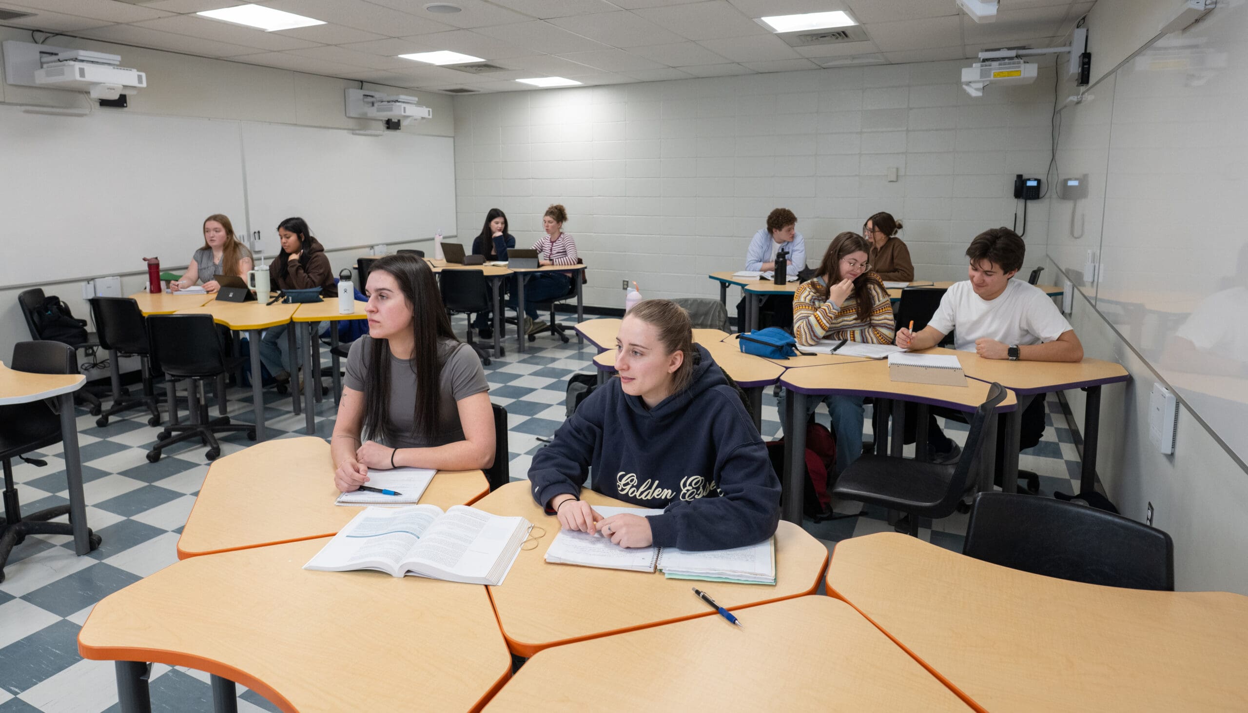 Les élèves sont assis à leurs pupitres dans une salle de classe ; certains écrivent ou lisent, leurs cahiers et manuels ouverts. La salle est dotée d'un sol en damier, de murs blancs et de projecteurs fixés au plafond. L'ambiance semble studieuse et concentrée.