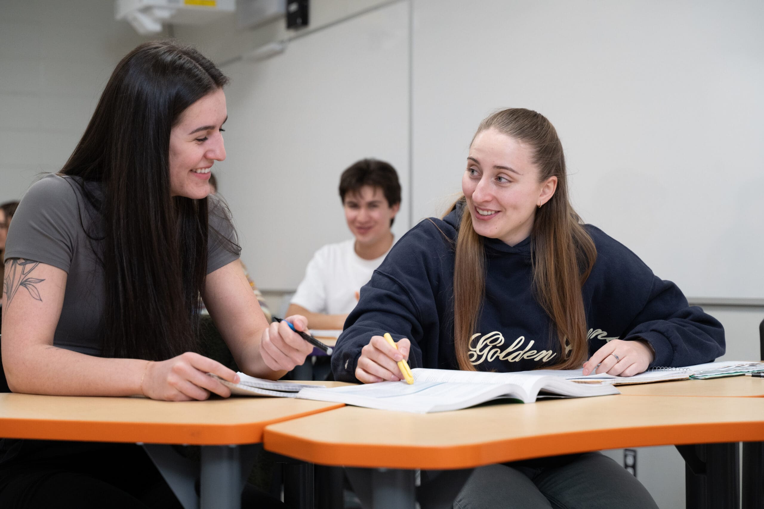 Deux élèves sont assis à leur pupitre ; ils sourient et travaillent ensemble sur leurs devoirs, munis de cahiers et de surligneurs. Un autre élève est assis et sourit à l'arrière-plan ; la scène se déroule dans une salle de classe.